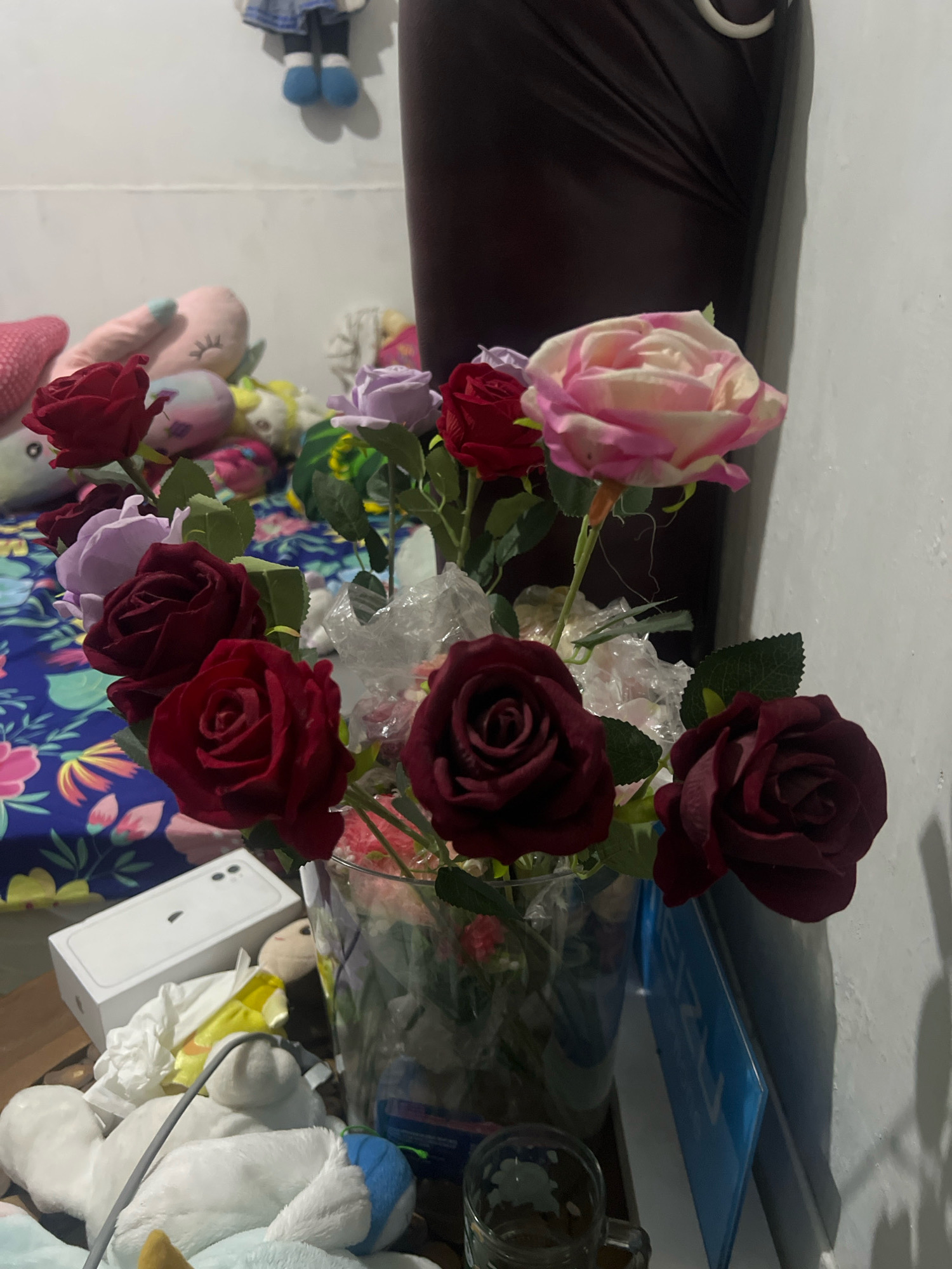 The image captures a close-up view of a bouquet of artificial roses in various shades of red and pink, displayed in a clear glass vase. The roses are arranged with some artificial leaves and clear plastic wrapping, giving a somewhat cluttered but decorative appearance. In the background, a portion of a dark brown headboard or piece of furniture is visible, along with a white wall.
The foreground of the image reveals a collection of stuffed animals and a white iPhone box, suggesting a domestic setting, likely a bedroom or living area. A gray cable extends across the stuffed animals, and part of a glass mug is visible on the right. The overall lighting appears to be dim, possibly from artificial light sources, casting soft shadows on the wall. The composition and content suggest an indoor scene in a personal space within Kecamatan Pati, Indonesia.