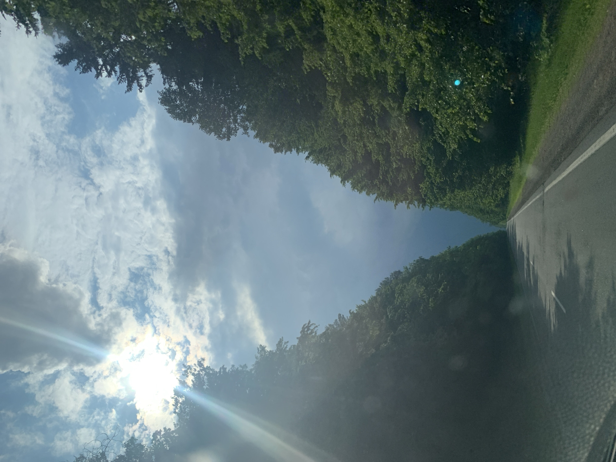The image shows a straight asphalt road with white dividing lines, lined on both sides by dense, green trees. The road recedes into the distance under a cloudy sky with bright sun rays piercing through the clouds on the left side. The sun's glare creates lens flare effects in the image. The environment suggests a rural or forested area, consistent with the location of Грозаўскі сельскі Савет, Belarus. There are no people or animals visible in the scene. The perspective suggests the photo was taken from a vehicle driving on the road.