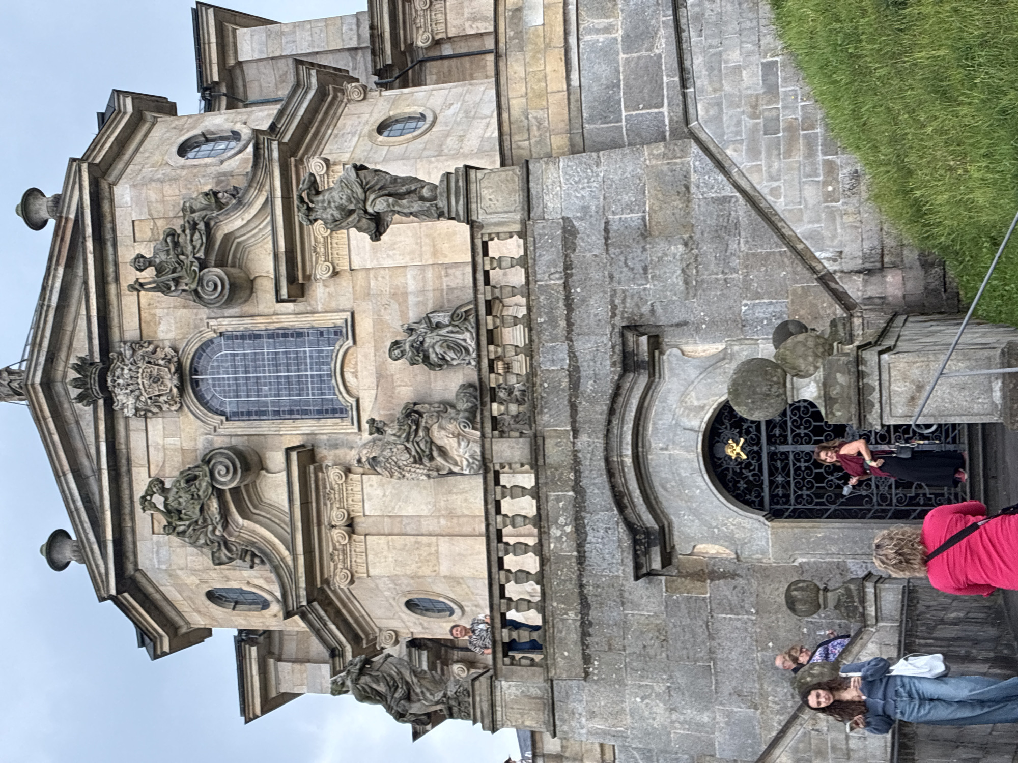 The image captures a low-angle view of the exterior of the Church of the Holy Trinity in Kuks, Czechia, a baroque hospital complex. The ornate sandstone facade features multiple levels adorned with sculptures and architectural details. In the foreground, several people are visible ascending and standing on the lower levels of the structure and stairs. One woman in a blue cardigan and jeans is looking at her phone, while another in a bright pink shirt stands with her back to the camera. A woman in a dark skirt and red top is paused on the steps near a decorative wrought-iron gate. Higher up, on a balcony, a person in a patterned shirt is leaning on the railing. The building itself is a grand stone structure with a central arched entryway at the bottom and multiple windows and sculpted figures on its upper sections. The sky is overcast, and a grassy slope is visible to the right of the building.