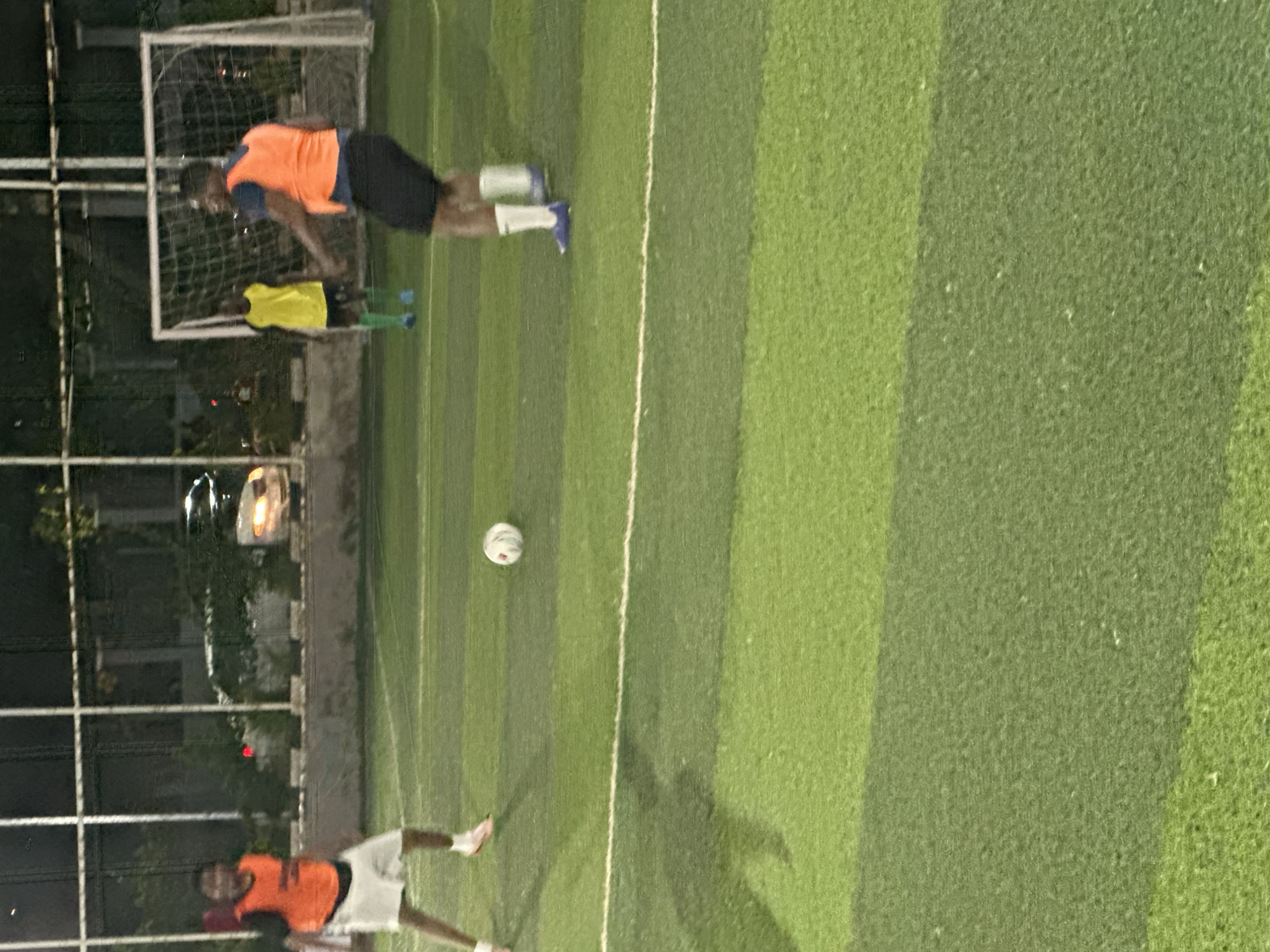 The image depicts a nighttime scene at a soccer field, likely in Abuja, Nigeria. Three individuals are visible, dressed in athletic wear. One person, on the left, wears a dark shirt with an orange vest and white shorts. Another person, on the right, is also wearing a dark shirt with an orange vest and black shorts, and is in the process of kicking a soccer ball. A third person, further back and in the center, is wearing a yellow shirt and green shorts. The field is artificial turf with visible white lines. In the background, there is a soccer goal net and fencing, with some trees and a blurry image of a car with illuminated headlights. The overall lighting suggests it is evening or night, with artificial lights illuminating the field.