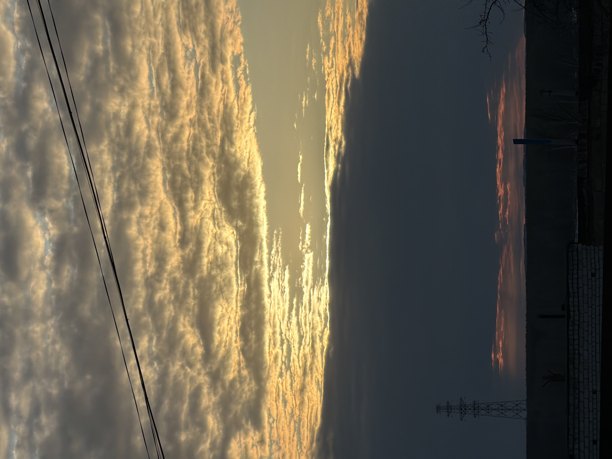 The image shows a sunset in what appears to be a residential area of Chaikovsky, Russia. The sky is divided between golden, textured clouds on the left and a dark, overcast sky on the right. Power lines are visible in the lower left corner. In the lower right, there is a brick building and a transmission tower silhouetted against the darkening sky. A sliver of orange-tinted clouds hints at the setting sun on the right.