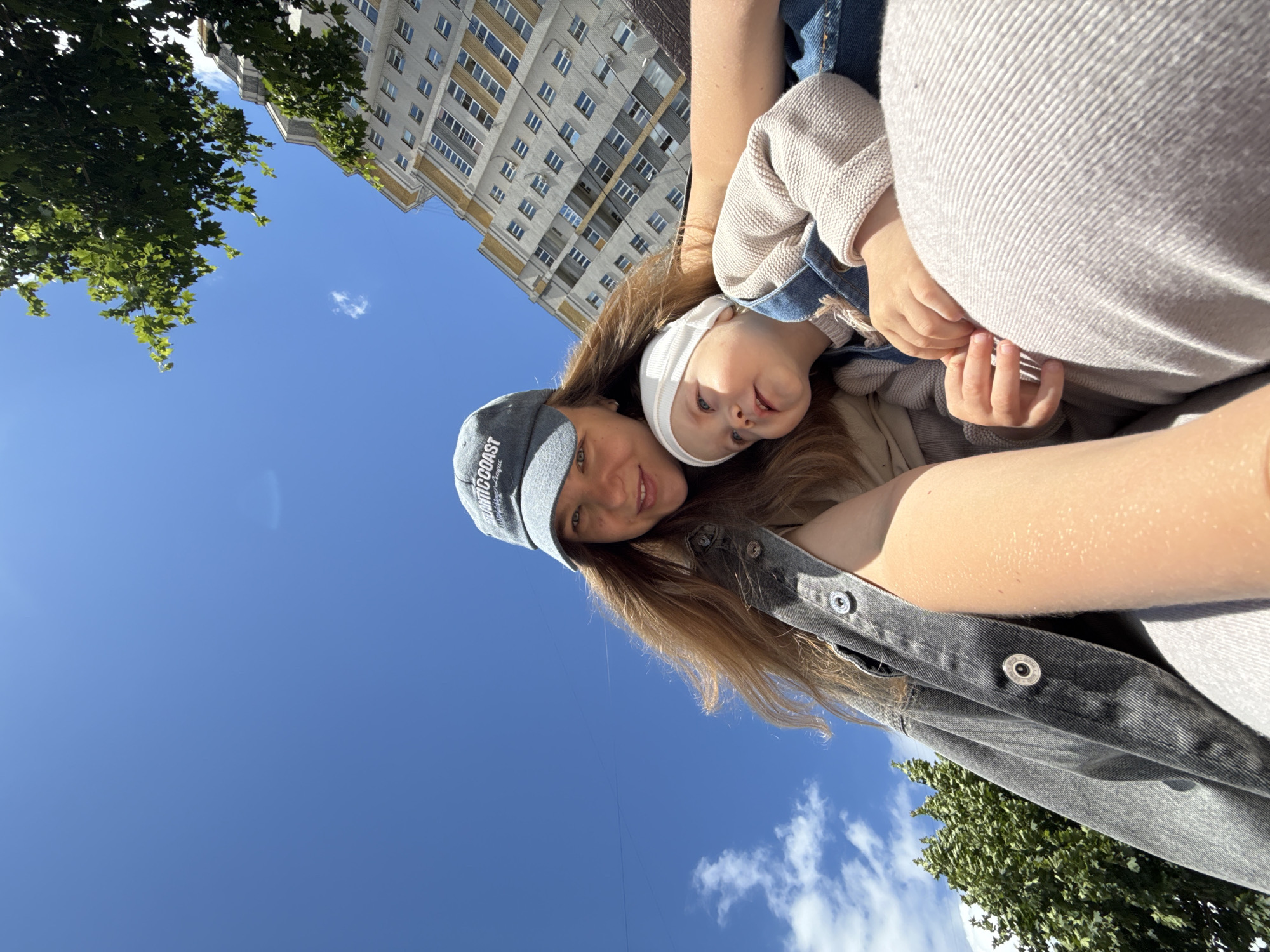 In a close-up, low-angle selfie taken outdoors, a woman and a baby are positioned in the foreground against a backdrop of a bright blue sky with scattered white clouds. The woman, wearing a gray baseball cap with the text "ATLANTIC COAST" visible, has long, light brown hair. She is smiling at the camera and wearing a dark gray denim jacket over a lighter colored top. To her right and slightly below her, a baby wearing a white cap and a light-colored knit sweater over denim overalls looks upward and to the side. The baby's hands are visible resting on the woman's lap.
In the mid-ground and background, a large, multi-story building with a light-colored facade and numerous windows dominates the right side of the image. Green trees with dense foliage are visible on the left and partially obscuring the top right of the building. The overall scene suggests an urban environment, possibly a city like Vladimir, Russia, on a sunny day.