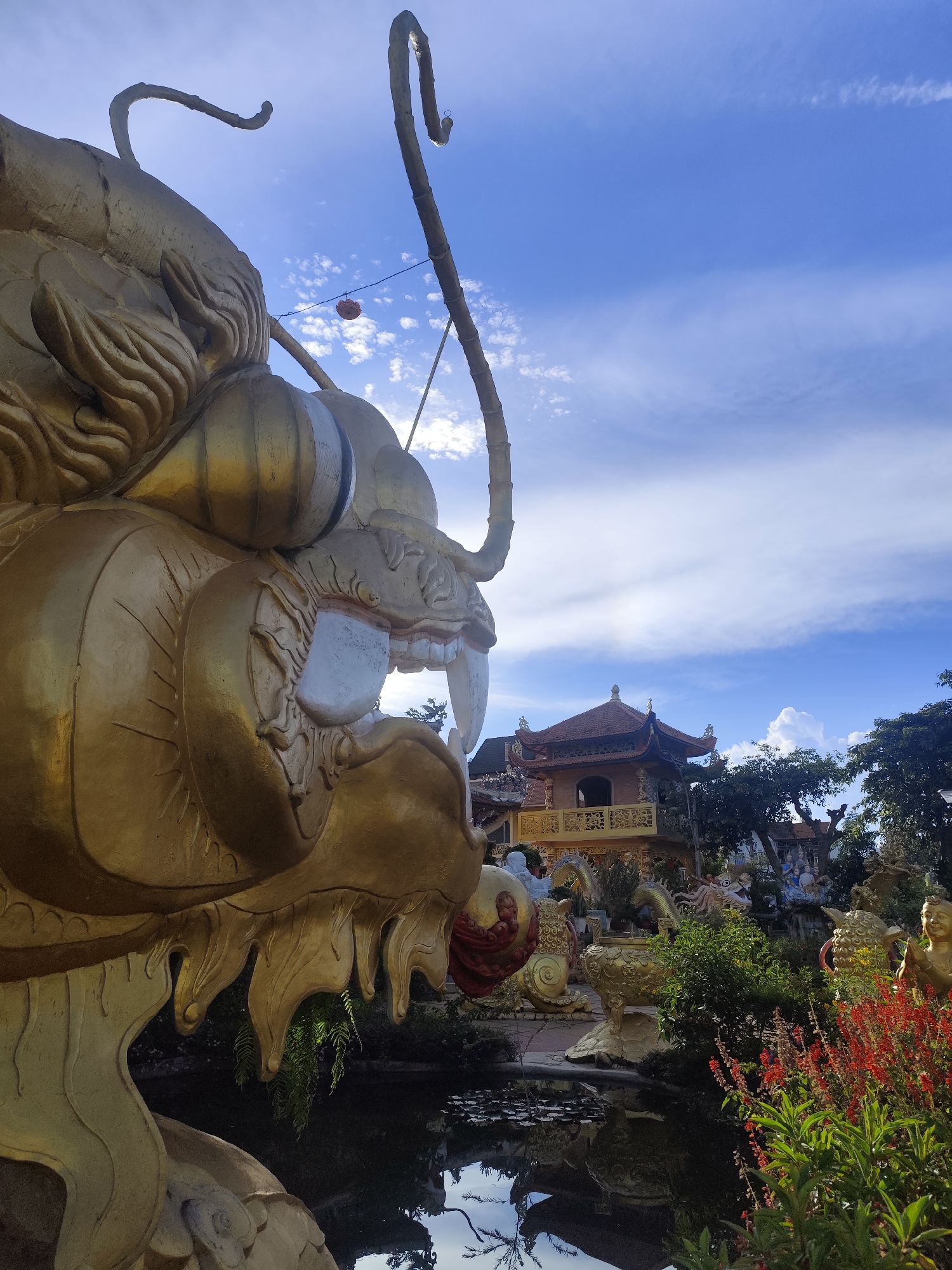 This is a ground-level, eye-level shot of a large, golden dragon statue, part of which is in the foreground and takes up a significant portion of the left side of the frame. The dragon statue is ornate, with detailed scales and sharp teeth visible in its open mouth. Behind the dragon, there is a pond reflecting the sky and surrounding structures.
Further back, a multi-tiered pagoda-style building with red roofing and yellow trim is visible. This building is surrounded by lush greenery, including trees and bushes with red flowers in the foreground on the right. Other statues, including smaller dragons and human figures, are scattered throughout the scene. The sky is a vibrant blue with scattered white clouds, suggesting a clear day.
This scene is located at the Linh Phuoc Pagoda, a Buddhist temple in Da Lat, Vietnam. The pagoda is known for its intricate mosaics made from broken ceramic and glass, and the grounds are adorned with numerous statues, particularly dragons. The presence of the large dragon statue in the foreground is a common feature of the pagoda's exterior. There are no discernible people actively engaged in any actions or interactions within the frame, though they may be present out of view or in the background.
