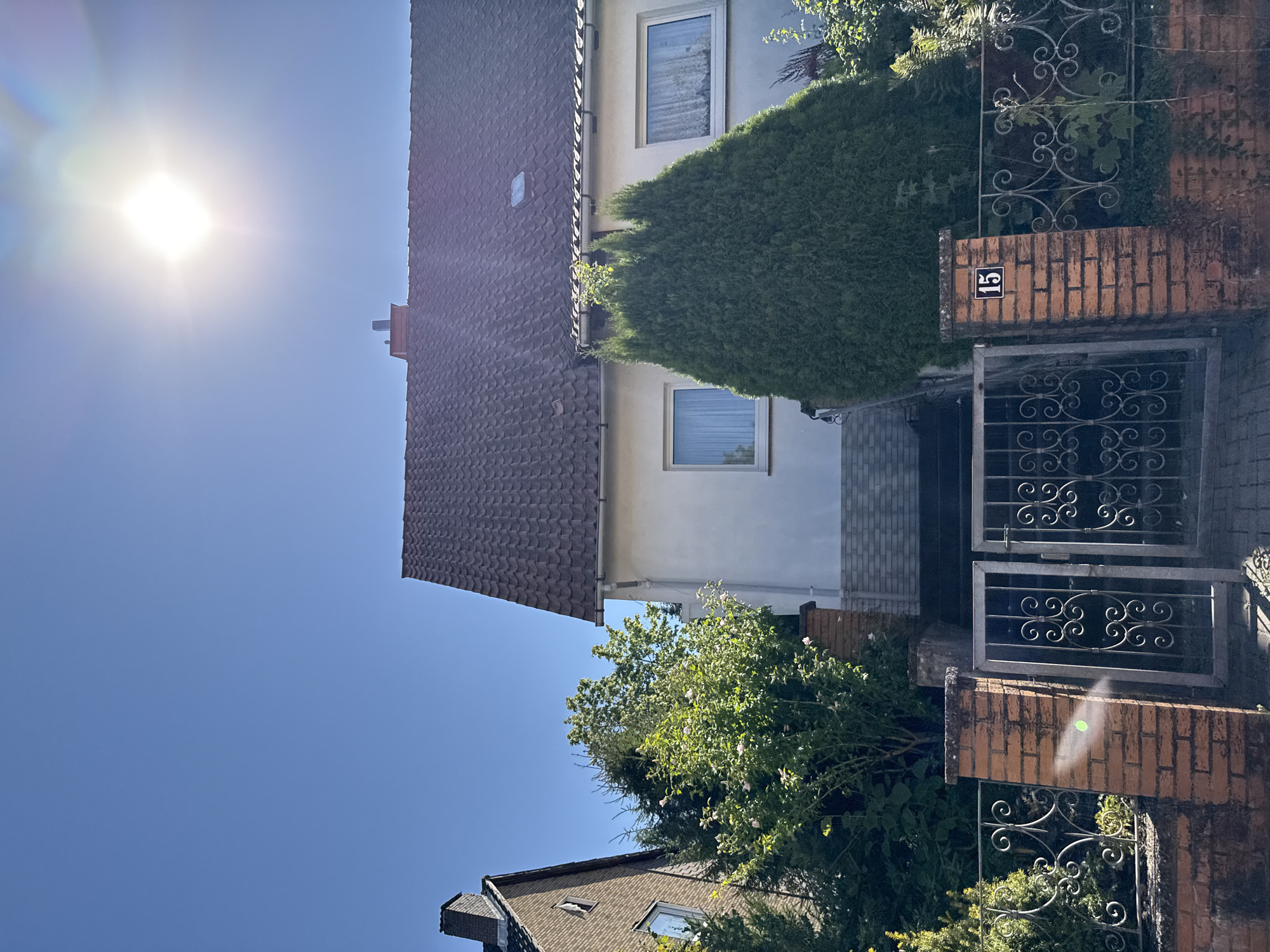 The image depicts a residential house on a sunny day in the city of Bad Sachsa, Germany. The house has a tiled roof and white exterior walls. In front of the house, there is a brick fence with a metal gate. A house number "15" is visible on one of the fence posts. Lush green trees and bushes surround the property. The sky is clear and blue with the sun shining brightly, causing lens flare in the upper right corner of the image. The scene is static and appears to be an outdoor, daytime shot.