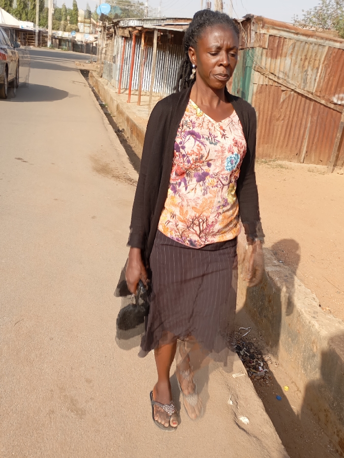 An adult female, appearing to be in her 40s or 50s, walks on a paved street in Jos, Nigeria, under bright sunlight. She has dark skin and black dreadlocks pulled back, wearing small dangle earrings. Her attire consists of a light-colored, V-neck top with a floral pattern in orange, purple, green, and blue, covered by a plain black cardigan. She wears a knee-length dark brown pinstriped skirt and black thong sandals with a decorative element on the right foot. She carries a dark-colored object in her left hand.
To her right, a concrete drainage ditch filled with debris runs parallel to the street, bordering an unpaved, reddish-brown dirt area. Beyond this, a series of single-story structures with heavily rusted corrugated metal walls and roofs, consistent with shops or stalls, line the street. In the background, on the left side of the street, a dark-colored vehicle is partially visible, with distant buildings and trees under a clear, bright sky. The street surface is light brown or tan, appearing dusty.