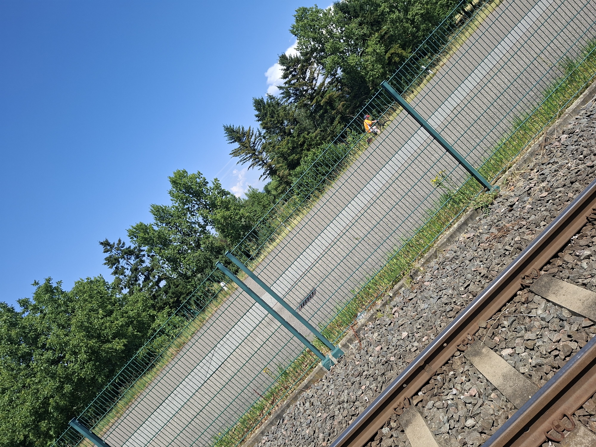 The image captures a sunny day scene near a railway line in Mannheim, Germany. The foreground features the gravel bed and metal rails of a railway track, angled diagonally across the frame. A green mesh fence runs parallel to the tracks, separating them from a paved path. Beyond the fence, lush green trees form a dense backdrop under a clear blue sky with scattered white clouds.
In the mid-ground, a person wearing a bright orange vest is visible on the paved path, walking away from the camera. The person appears to be engaged in some activity along the path. The time of day is clearly daytime, indicated by the bright sunlight and clear sky, suggesting fair weather. The overall mood is one of an ordinary day in an outdoor setting. No visible text is present in the image.