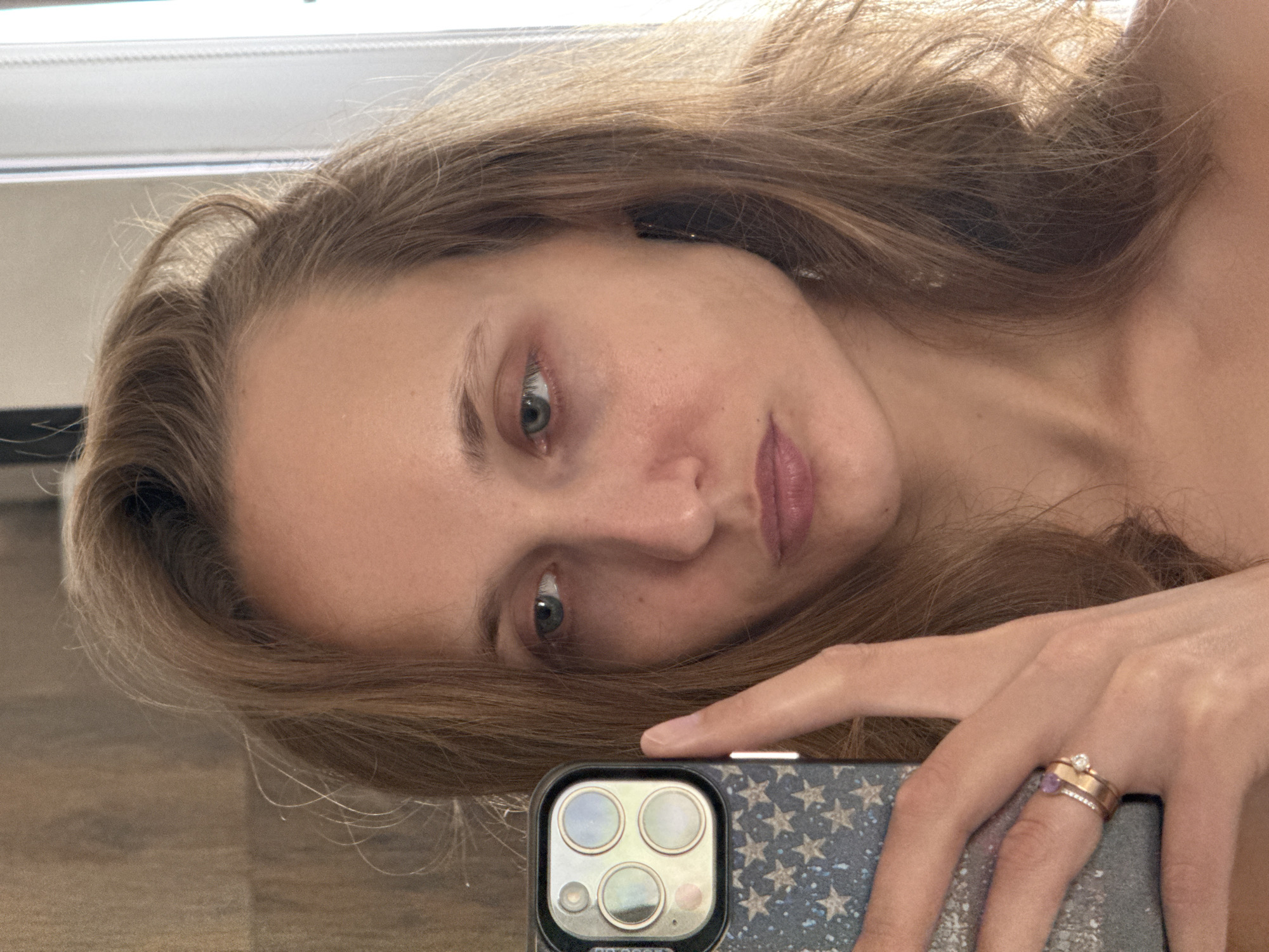 A medium close-up selfie, taken indoors, shows a woman with light brown hair, holding a phone in her left hand to take the picture. Her gaze is directed towards the phone screen, her lips slightly parted. She is wearing two rings on her left ring finger. The background suggests an indoor setting with a wooden-textured wall and a bright window frame on the right. There is no indication of the city of Подшивалово, Russia, or any specific urban features.