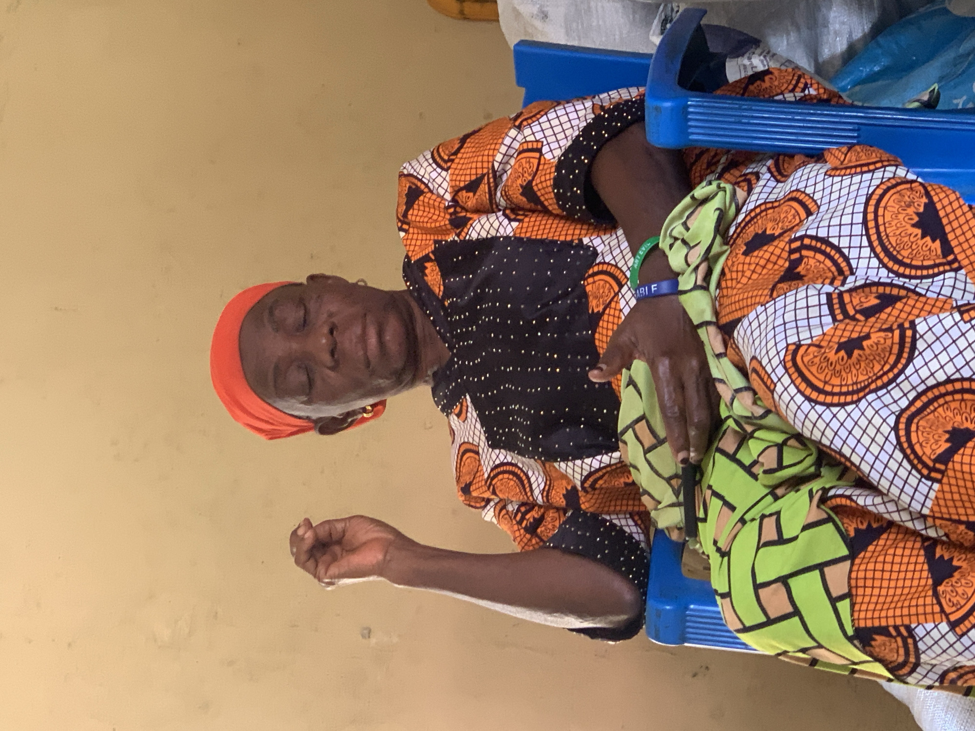 A medium shot shows an elderly Black woman wearing a vibrant orange headscarf and a colorful patterned dress, seated in a blue plastic chair against a plain beige wall. Her eyes are closed, and her head is tilted slightly down. Her right hand is raised, with her index finger and thumb touching, in a gesture that could indicate contemplation or prayer. She is holding a folded green and orange fabric in her lap. A green beaded bracelet is visible on her left wrist. The scene appears to be indoors, possibly in a humble dwelling or community center, given the simplicity of the furnishings and the textured, slightly worn appearance of the wall. The overall impression is one of quiet repose or reflection. The location is the city of Jos, Nigeria.
