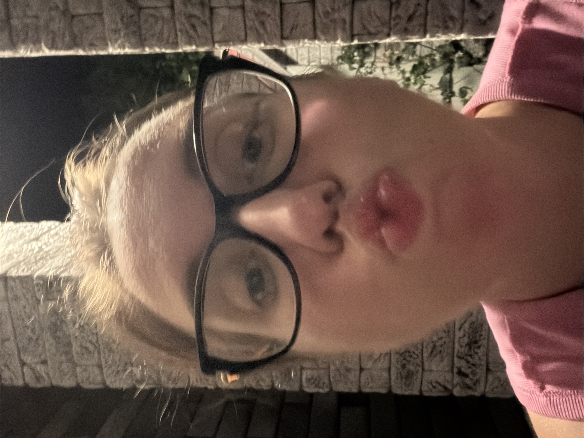 A woman wearing glasses and a pink shirt poses for a selfie in front of a brick wall at night. Her lips are pursed. The background is dark, with some foliage visible behind the wall. The location is identified as Bila Tserkva, Ukraine.