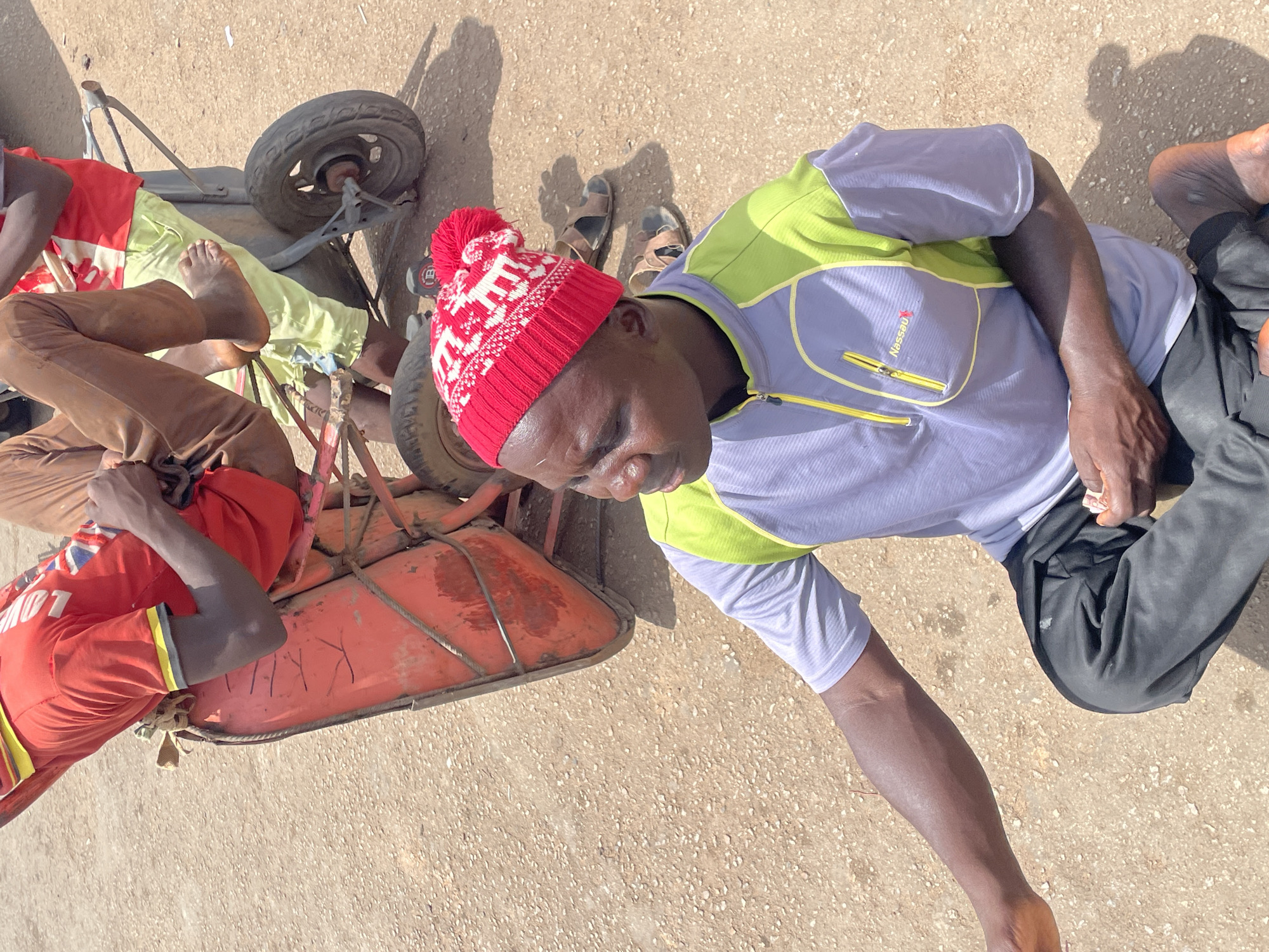 The image, likely taken in Jos, Nigeria, shows two individuals near a low, three-wheeled cart. One man wears a red and white knit hat, a light blue and green jacket, and black pants. He is barefoot and partially kneeling on the ground. The other man, partially visible, is in a red shirt with yellow trim and brown pants, lying in the cart. The cart itself is painted red and appears weathered, with words etched on the side. The surface is rough, likely concrete or a similar material. The lighting suggests it is daytime.