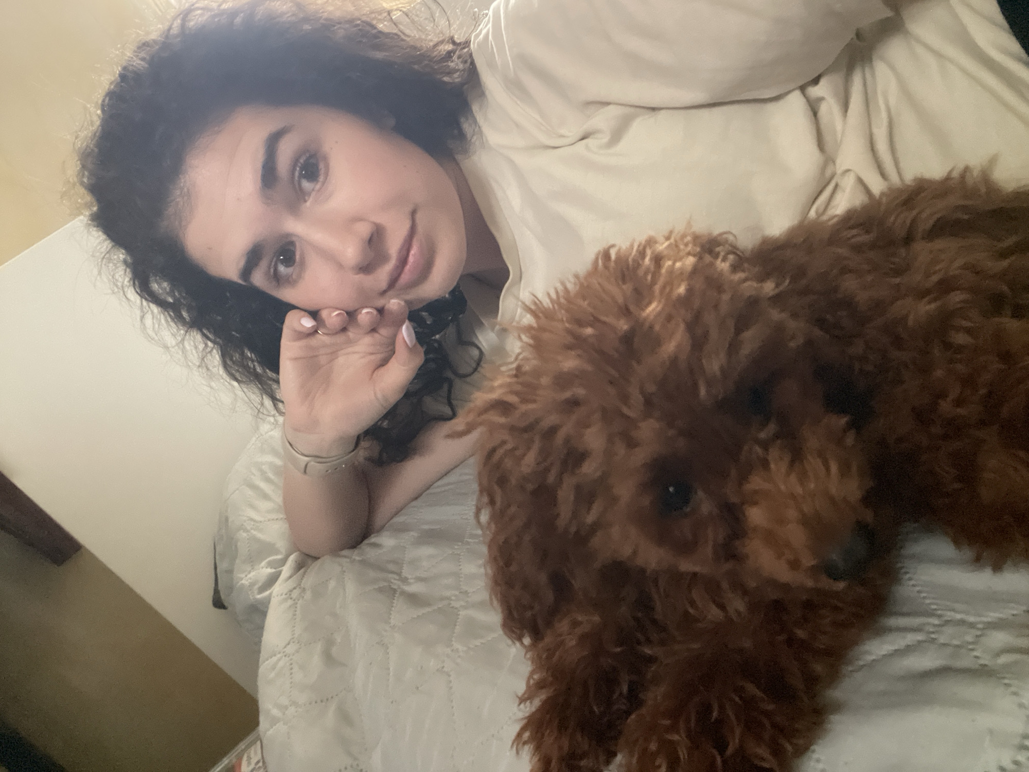 A woman with curly dark hair lies on a quilted, light-colored bedspread, resting her face in her hand. She is looking towards the camera. Next to her lies a brown, curly-haired dog. The background shows a light-colored wall and bedding. There is no information in the image indicating its location in Sofia, Bulgaria, or any other specific location.