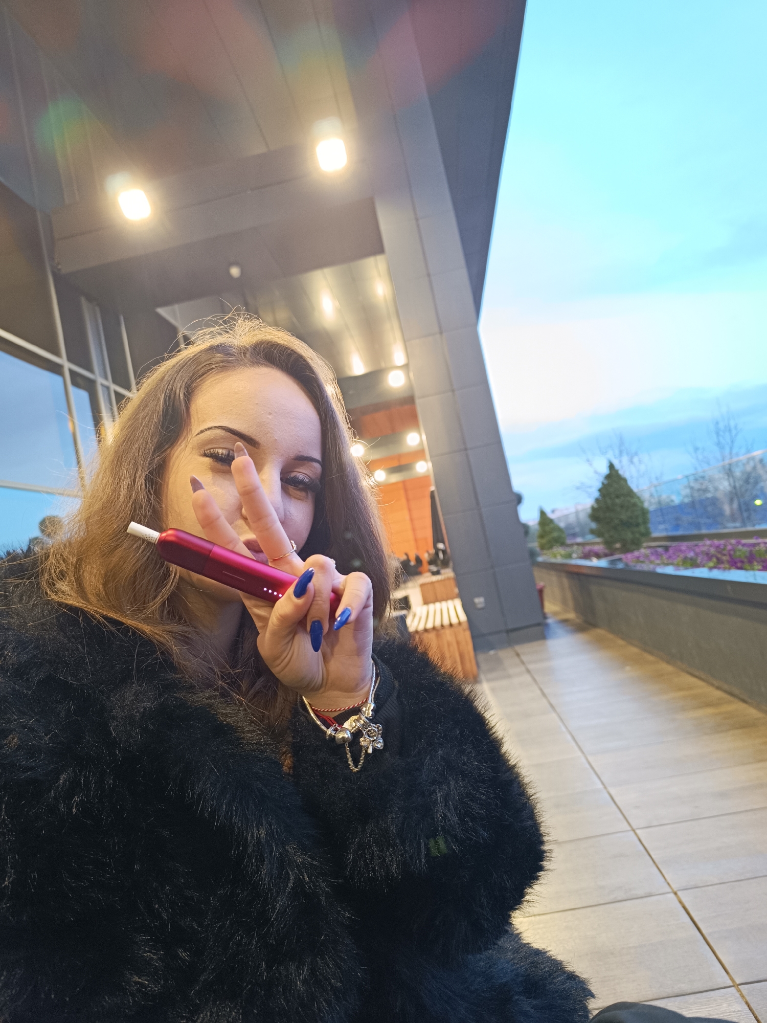 A young woman is posing for a selfie. She is holding a red electronic cigarette or heat-not-burn device in her right hand, making a peace sign with her fingers. She has long brown hair, dark eye makeup, and manicured nails painted a bright blue. She is wearing a black fur coat and a charm bracelet.
The background shows an outdoor area, possibly a terrace or balcony. There is a modern-style building with large windows and gray cladding. In the distance, there are trees, landscaping with purple flowers, and a cityscape. The sky is a light blue color.
While I cannot pinpoint the exact location in Plovdiv without more information, the modern architecture and terrace setting suggest a possibly modern hotel, business or residential area.