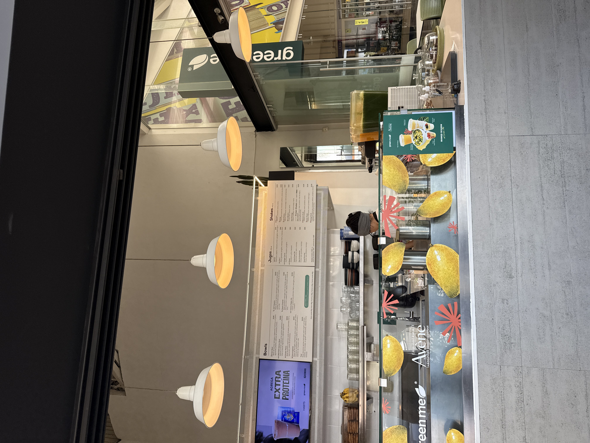 Here is a description of the image:
This is an interior shot of a Green Me Avene store, likely in Puebla, Mexico, based on the presence of the words "gree", "extra proteína" and "Jugos" in Spanish on the menu and promotional materials. The store appears to specialize in healthy foods and beverages like bowls, juices, and shakes.
In the image, a menu board is on the back wall, displaying the items offered. A worker wearing a head covering is positioned behind the counter, likely preparing an order. Clear cups are stacked on a shelf behind the employee. A refrigerated display case with mango-themed decorations along the front is visible, and the wall to the right of the display case is light gray. Hanging from the ceiling are several modern, rounded lamps. The store has glass windows allowing a view of the mall interior. A black frame is seen on the left side of the image and along the top where the store logo is.