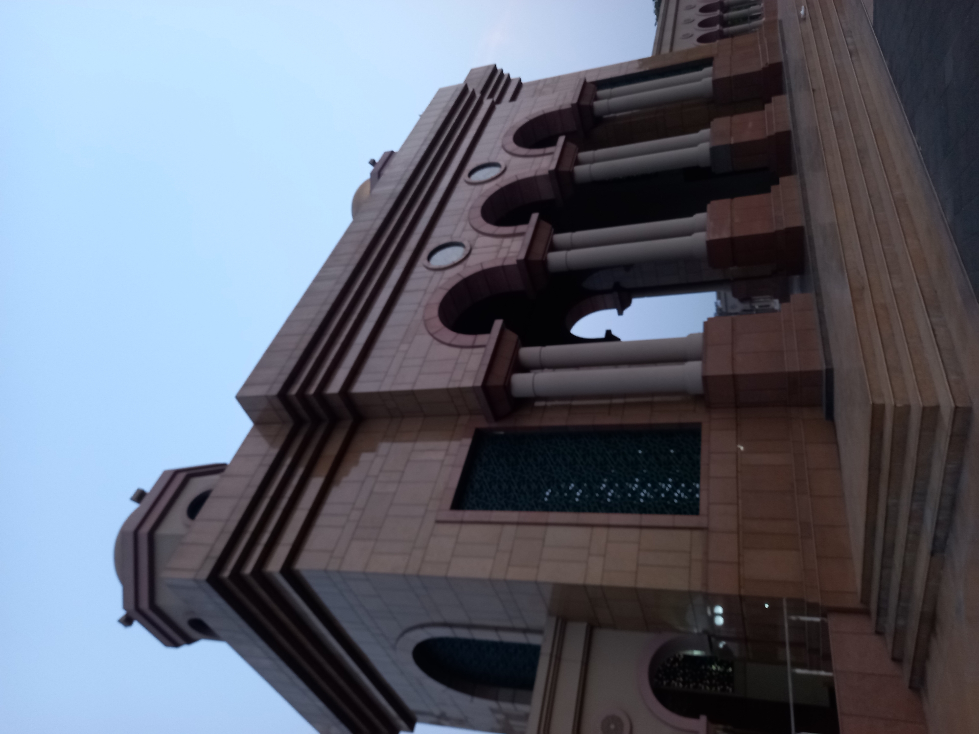 The image shows the exterior of the Islamic Center of Samarinda, Indonesia, also known as Masjid Raya Darussalam. The building features a grand architectural design with a central dome and multiple minarets. The facade is adorned with intricate patterns and calligraphy, characteristic of Islamic art. The mosque is surrounded by a well-maintained courtyard and a serene atmosphere, with lush greenery visible in the background. The sky is clear and blue, suggesting a pleasant day. The mosque is a prominent landmark in Samarinda, attracting visitors and worshippers from all over the region.