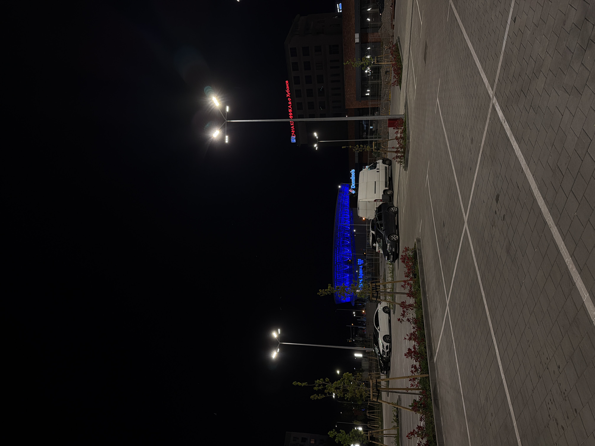The image shows a parking lot in Burgas, Bulgaria, at night. Several vehicles are parked in designated spaces. A building with illuminated signage in Cyrillic is visible in the background. A Domino's delivery van is also present. A large building or arena is lit up with blue lights. Streetlights illuminate the parking area. The ground is paved with patterned stones, with clearly marked parking lines. Small trees are planted along the edge of the parking lot. The sky is dark and clear.