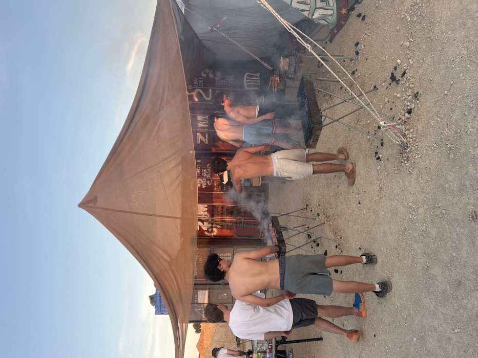 The image shows a group of five men gathered beneath a large canopy in Tze'elim, Israel. The men are standing around several charcoal grills, cooking and smoking. The setting is a desert landscape. Two of the men are wearing shorts and flip-flops, while the others are shirtless and wearing shorts or swim trunks. One of the men is holding a grill spatula. The canopy is made of brown canvas and is supported by metal poles. In the background, a metal container labeled with the word “ZIM” is partially visible. The ground is composed of gravel and sand. The sky is clear and blue. The time appears to be early evening, as the light is warm and the shadows are long. The scene evokes a sense of relaxation and camaraderie. The men are enjoying the outdoors and the process of cooking together.