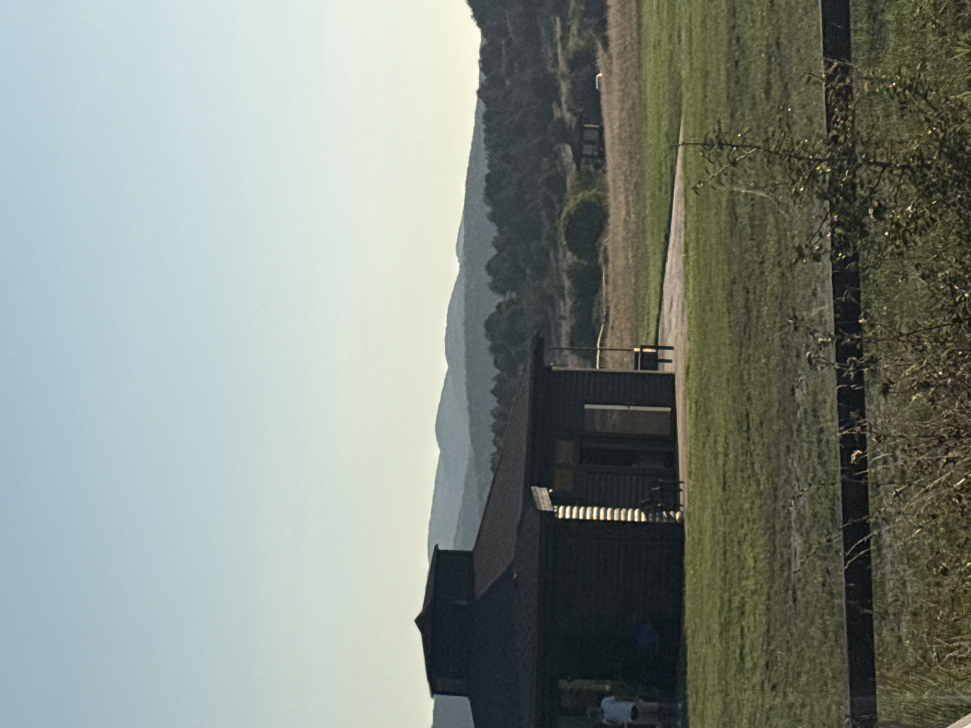 The image captures a view of a small, dark-colored building with a multi-level, octagonal roof. The building is situated in a grassy, open area with a paved walkway leading up to its entrance. In the background, rolling hills covered in trees recede into the distance under a clear, pale blue sky. A lone figure in a white shirt and shorts stands to the left of the building. In the far distance, another small, open-sided structure is visible amidst the trees. The foreground is dominated by a wooden fence and dry vegetation. The scene appears to be set in a rural or park-like environment.