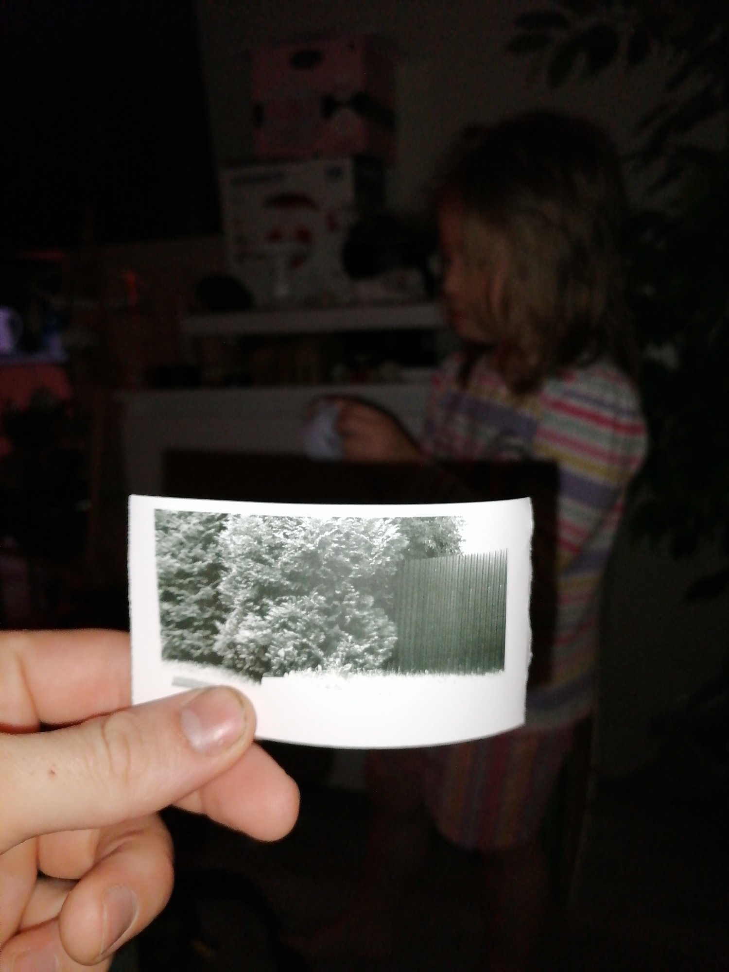 An adult's hand is holding a black and white photograph that depicts a green wooden fence and some leafy trees. Behind the hand and photograph, a young child wearing a striped shirt is visible, partially out of focus. The background is dark and indistinct, suggesting an indoor environment with low lighting.