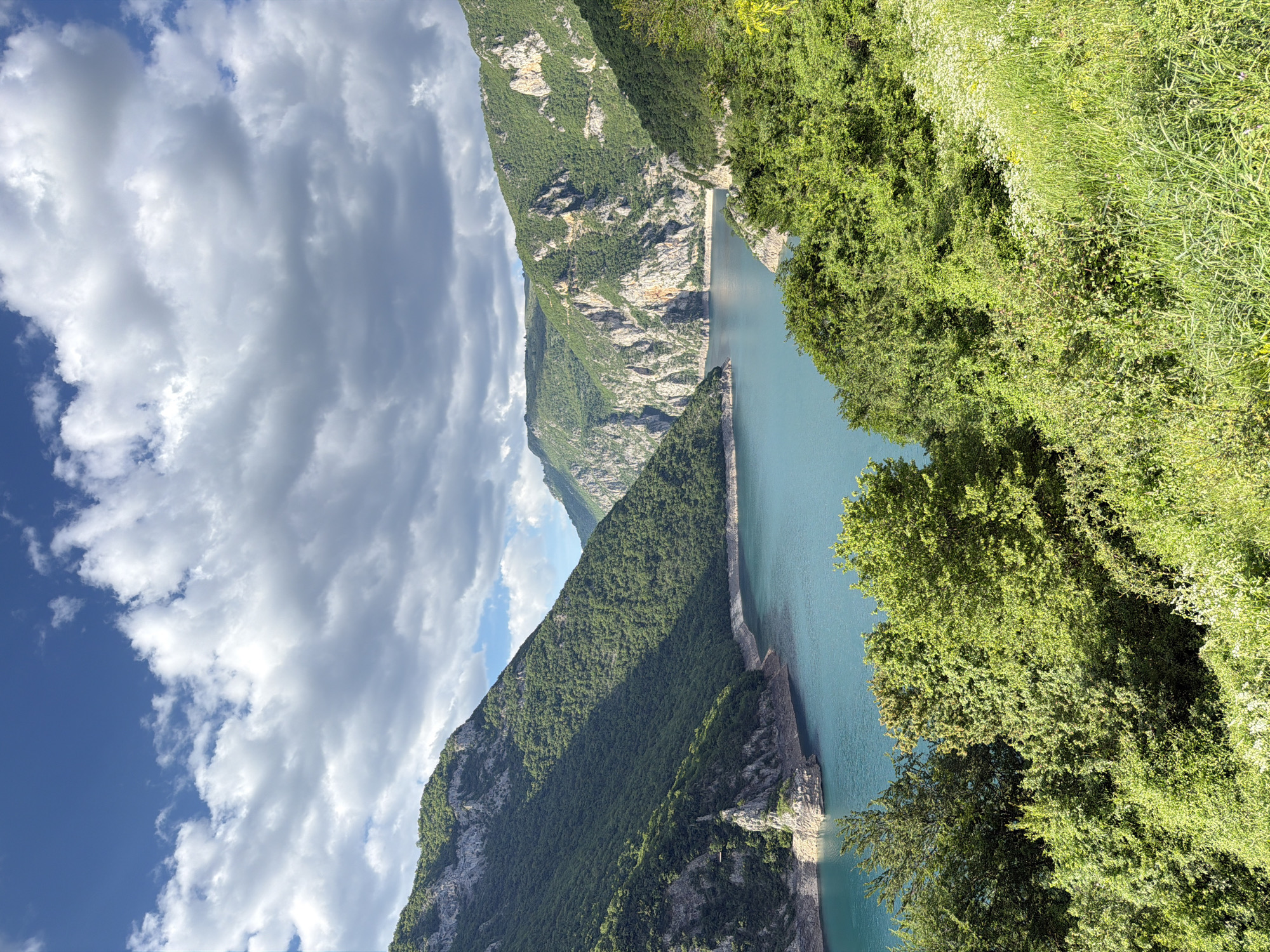 The image shows a natural landscape with a long, narrow body of water, likely a lake or river, situated between steep, green mountains. The mountains are predominantly covered with vegetation, with some areas displaying bare rock formations. On the right side, there are trees and dense foliage, obscuring some of the water's edge. The sky overhead features a combination of blue patches and cumulus clouds. The image is taken from a high vantage point, looking down onto the water and the surrounding landscape. The location is the city of Plužine, Montenegro.
