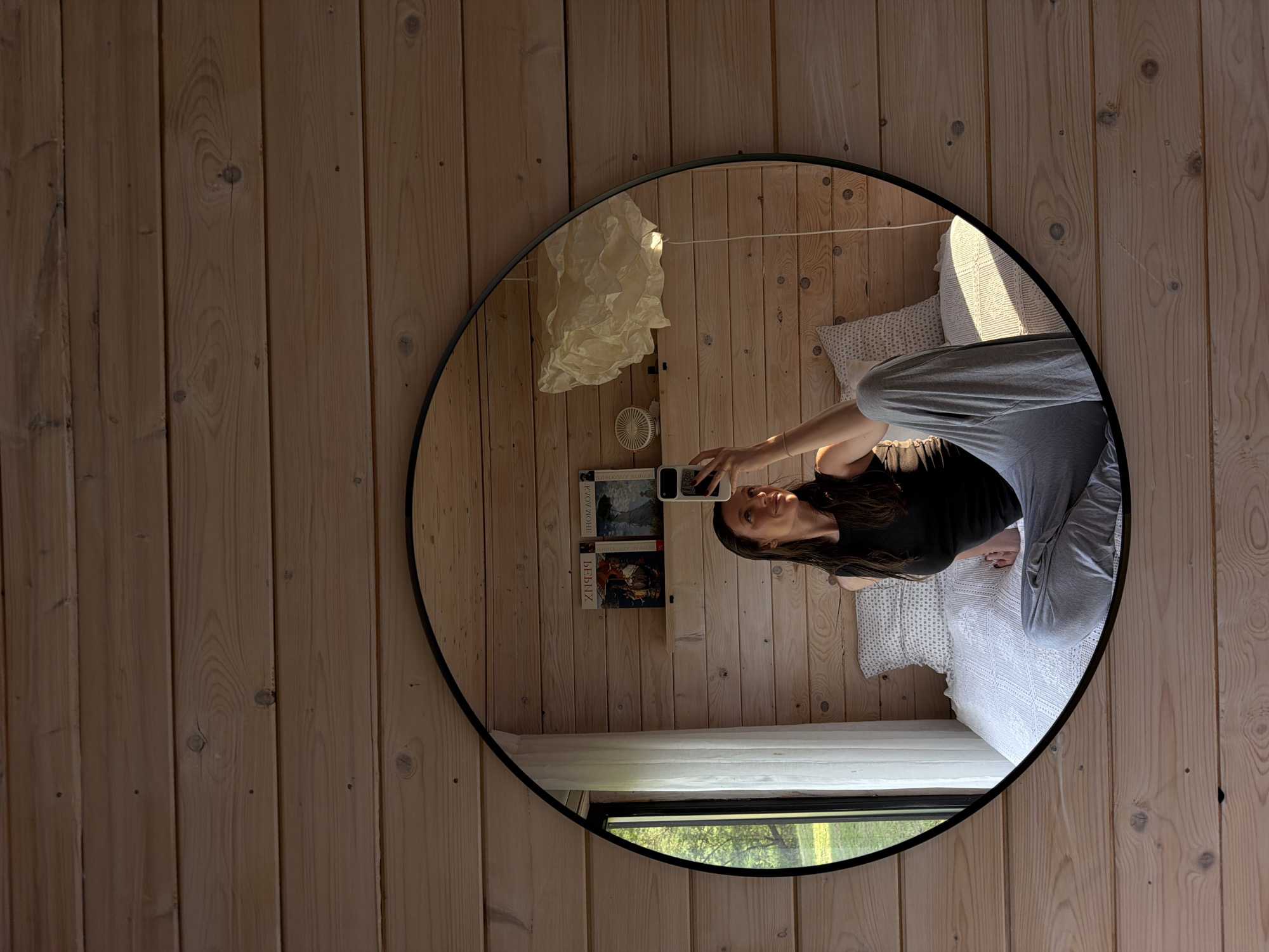 A woman takes a mirror selfie in a room with wood paneling. The reflection reveals a bed with white patterned bedding, a shelf with books, a window with a green view outside, and a paper light fixture. The woman is wearing a black top and grey sweatpants, holding a phone up to take the picture. There is no information about Prudki, Russia in the image.