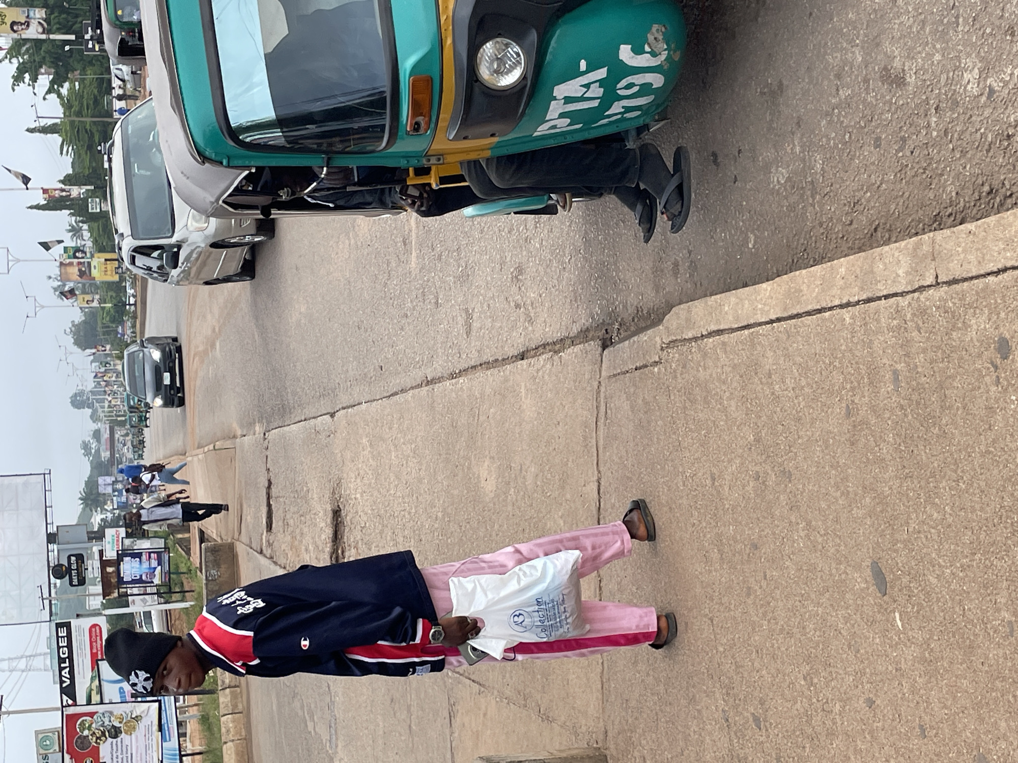 The image shows a street scene in Jos, Nigeria. In the foreground, a young man wearing a black beanie, a navy blue jacket with red and white stripes, and pink pants stands on a sidewalk, holding a white plastic bag. To his right, a turquoise auto-rickshaw, identified by the inscription "PTA- 796," is parked by the roadside, with two men visible inside. Further down the street, other vehicles, including a silver SUV and a black car, are in motion. The environment appears to be a commercial area with various billboards and signs visible in the background, suggesting a busy urban setting. The sky is overcast, indicating a cloudy day.