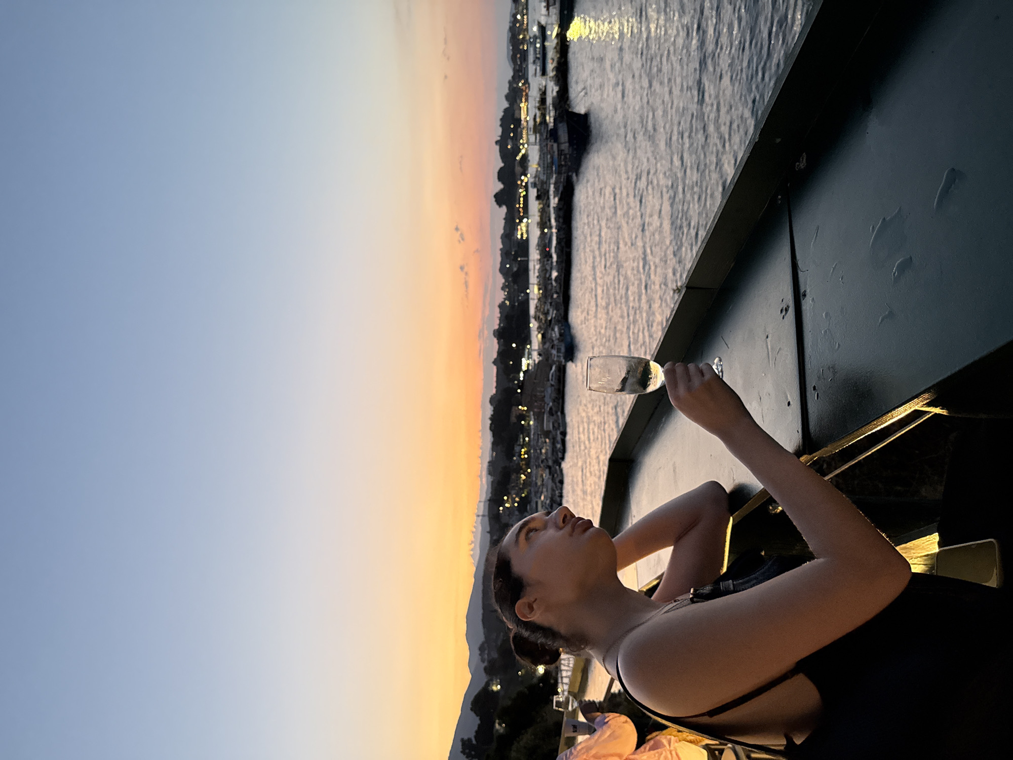 A young woman is seen from the chest up, holding a wine glass and looking out at a harbor during sunset. The woman has dark hair pulled back into a bun. She is wearing a black tank top. The setting appears to be a waterfront establishment, possibly a restaurant or bar, in Laganas Municipal Unit, Greece, given the presence of docked boats and a coastal landscape. The sky is a gradient of blue and orange, indicative of twilight. The water reflects the soft light of the sunset and the lights from the shore. The overall mood is relaxed and scenic.