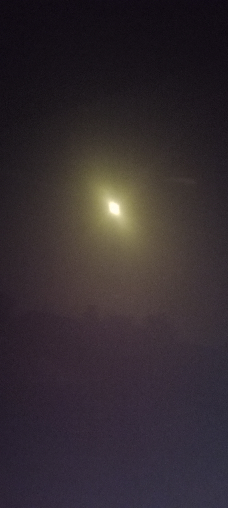 A vertical, wide shot captures a hazy night sky above Adai, Spain. The scene is dominated by the dark tones of the night, with a subtle gradient moving from a lighter yellow hue near the top to a deep purple at the bottom. In the upper half of the frame, a bright, diamond-shaped light source is visible, haloed by a soft, yellow glow. This light appears to be a streetlight or a similar artificial illumination, its intensity diffused by the atmospheric haze. The lower half of the image is a darker expanse, with the hazy atmospheric conditions obscuring any ground-level details or significant features within the city of Adai. No people or specific objects are clearly discernible.