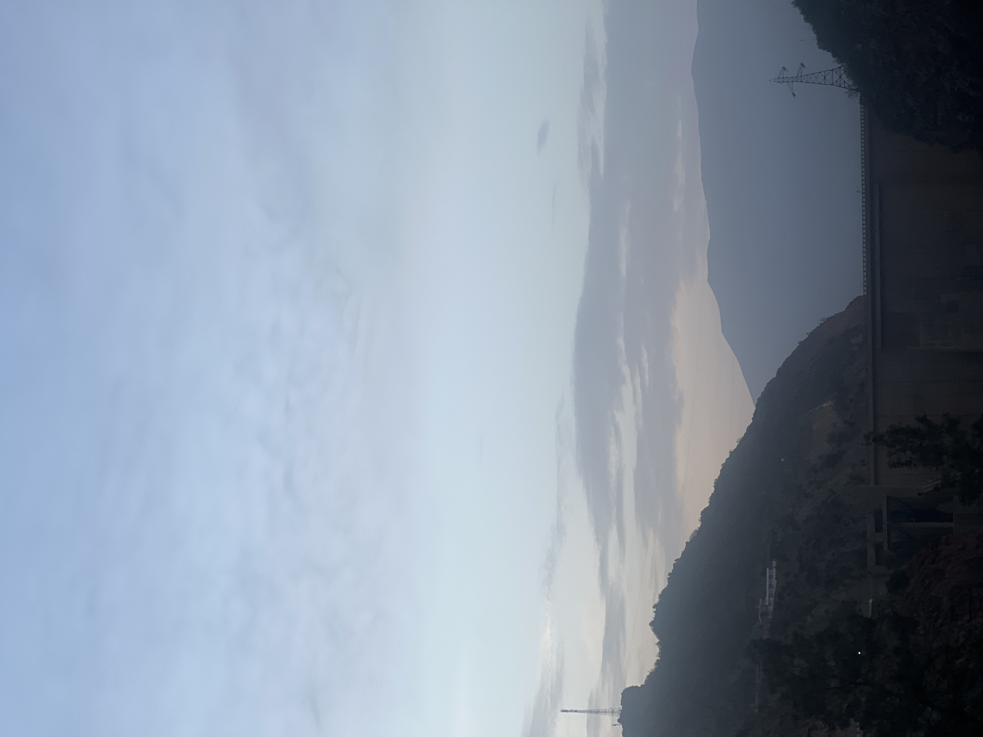 The image shows a landscape likely near Bin El Ouidane, Morocco. The right side of the image depicts a mountain range and a dam structure. A power line transmission tower is visible on the top right. The left side is dominated by a sky with clouds. The light suggests it's either early morning or late afternoon. The image seems to be taken from ground level, facing toward the mountains.