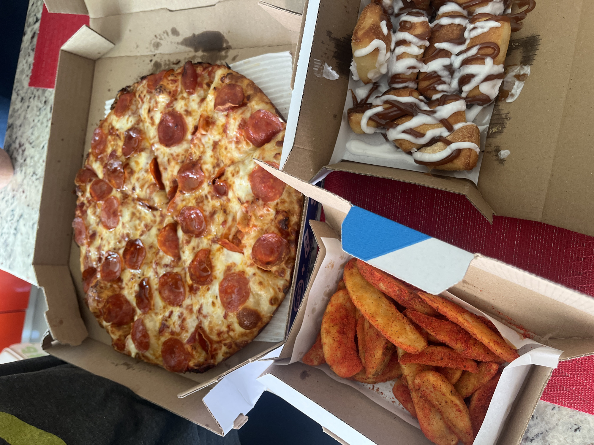 The image shows a collection of takeout food items, likely from a pizza chain, arranged in their respective cardboard containers. A pepperoni pizza occupies the left-most box, with its circular slices visible. In the top right box, there are pastry rolls drizzled with white and brown icing. In the bottom right box, a serving of potato wedges seasoned with a reddish-orange spice blend is visible. The boxes appear to be placed on a surface that includes a red textured placemat or tablecloth. The location in the city of Barrio de Nuevo León, Mexico cannot be confirmed from the provided image.