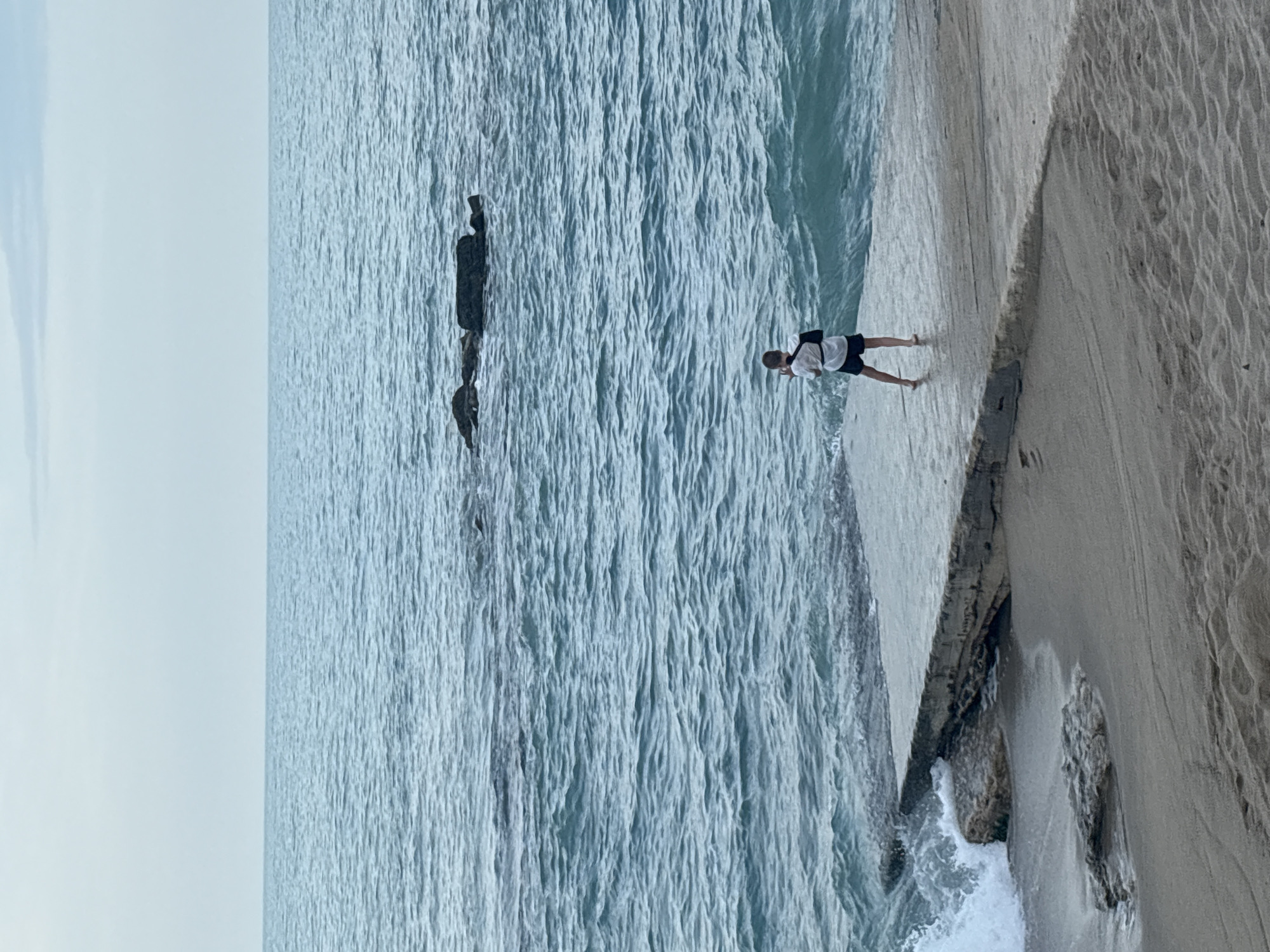 An outdoor, slightly elevated shot captures a coastal scene on a clear day. The upper half of the image is dominated by the calm, light blue waters of an ocean or large body of water, marked by subtle ripples and small waves. The sky, a paler shade of blue and mostly clear, occupies the top portion of the frame, suggesting daytime.
In the middle ground, partially submerged dark, weathered structures resembling rocks or old pillars rise from the water, creating a point of interest amidst the expanse of blue.
On the right side of the frame, a concrete pathway or barrier runs along the water's edge, transitioning into a sandy beach with visible texture from wind or waves. A lone individual, appearing to be male, stands barefoot on this concrete structure. He wears a light-colored t-shirt and dark shorts, with a small dark bag slung over his shoulder. He is positioned facing slightly away from the camera, seemingly looking out towards the water and the horizon. His stance appears relaxed, suggesting he is observing the scene or enjoying the coastal environment. Small white splashes of water indicate gentle waves lapping against the concrete edge.
The overall atmosphere is tranquil and serene. There is no visible text in the image.