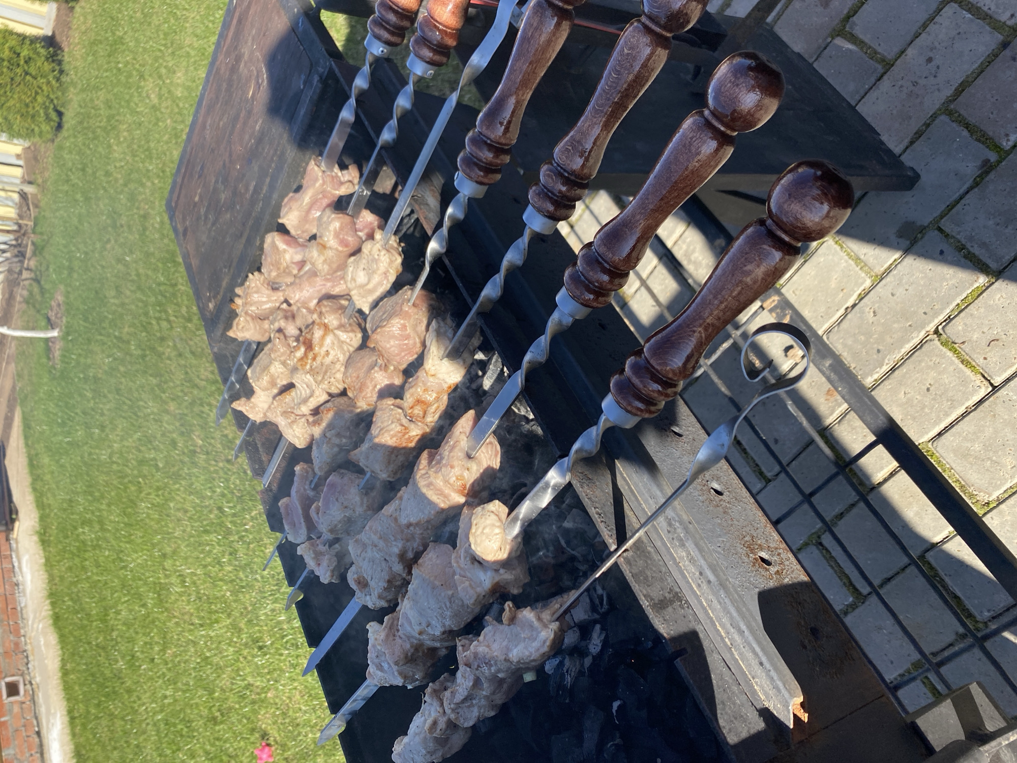 The image shows skewers of meat cooking on a barbecue grill in an outdoor setting. The location is Гаревая, Russia. The skewers have wooden handles and are filled with chunks of meat. Smoke rises from the barbecue. The background consists of grass and paving stones.