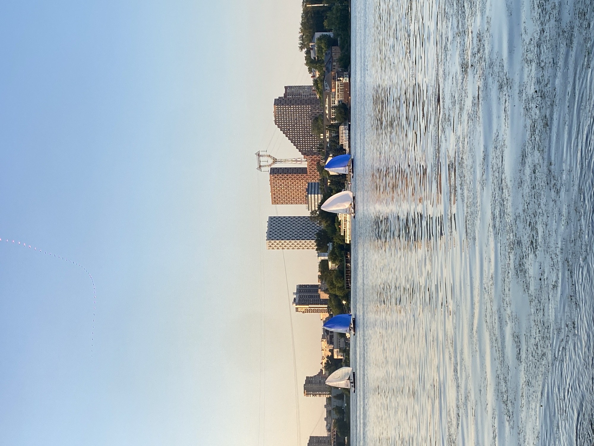 The image depicts a serene waterfront scene in Khimki, Russia, with the city's skyline forming a backdrop. In the foreground, a body of water reflects the soft hues of the sky and the distant buildings. Several sailboats are gliding across the water, their sails catching the gentle breeze. The sailboats are white and blue. The city skyline is characterized by a variety of buildings, including modern high-rises and older structures, interspersed with trees and vegetation. The overall atmosphere is peaceful and tranquil, suggesting a calm evening or morning by the water. The sky is clear, with a subtle gradient of blue and a hint of warm light, possibly from a rising or setting sun. There are no people visible in the image, only the sailboats as the primary subjects on the water. The scene appears to be a typical representation of urban waterfront life, where nature and cityscape coexist harmoniously.