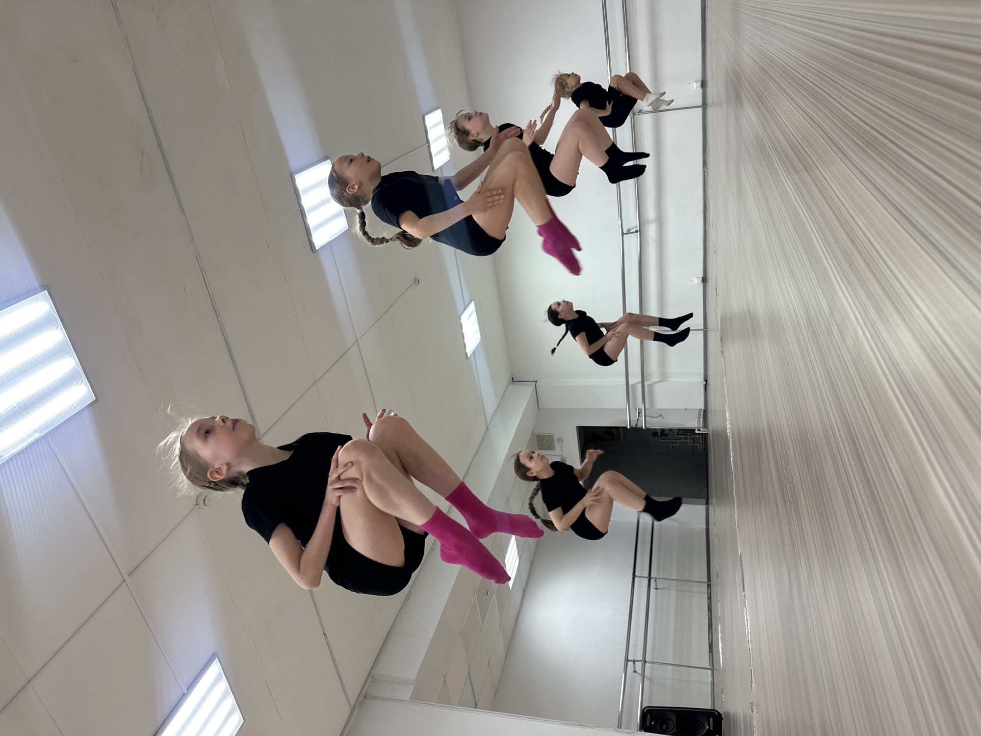 The image shows a group of girls in a studio, apparently practicing a choreographed routine or gymnastic exercises in front of a mirror. The girls are wearing dark athletic clothing and socks (some pink, some black). They are positioned in a line, facing the mirror, with their legs bent and arms around their knees. The room has a tiled ceiling with fluorescent lighting. The wall with the mirror is partially covered with vertical lines. The scene appears to be a dance or gymnastics studio based on the mirrored wall, the presence of bars near the mirror, and the activities taking place. The image suggests that the location is the city of Votkinsk, Russia.