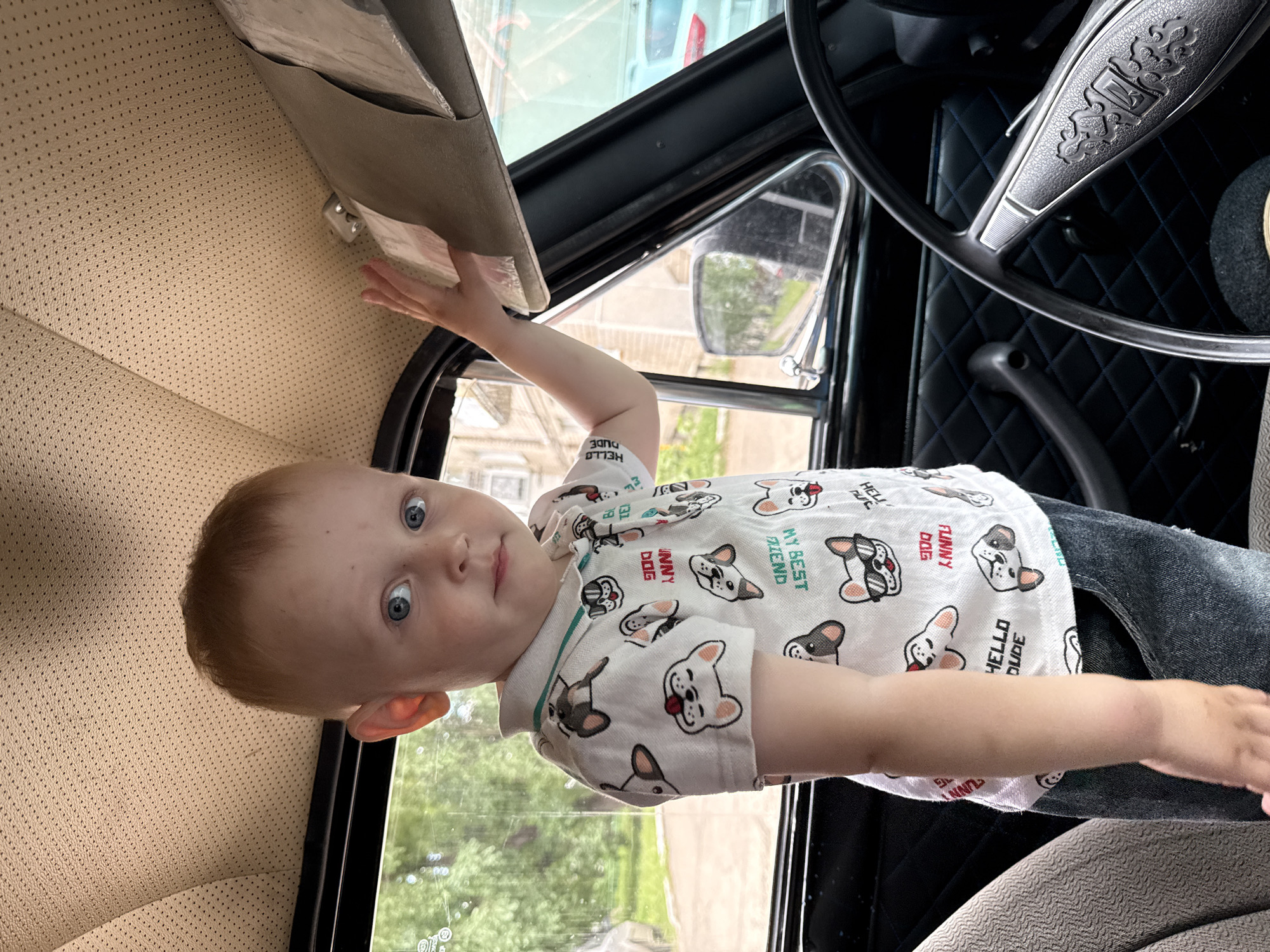 A young Caucasian child with fair skin and blue eyes stands inside a car, looking directly at the camera. The child has short, light brown hair and is wearing a white polo shirt with a repeating pattern of cartoon dogs. The child's left hand is raised, touching the car's sun visor. The interior of the car is visible, including a beige perforated headliner, a black steering wheel, a black quilted door panel with a chrome handle, and a portion of the driver's seat. Outside the car window, trees and buildings are visible, suggesting an urban or suburban setting. The image appears to have been taken in Chaikovsky, Russia, based on the provided location context.