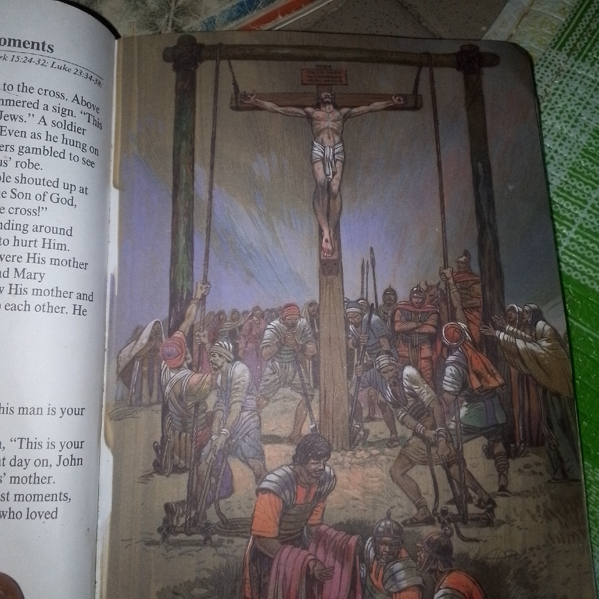The image depicts a scene of the crucifixion of Jesus Christ. In the foreground, a crowd of people, some dressed in Roman soldier attire and others in common attire, are gathered around the base of three crosses. One of the crosses is prominently displayed in the center, with Jesus crucified upon it. His body is emaciated, and he wears a simple loincloth. Above his head, a sign is affixed to the cross. The crowd below appears to be in various states of reaction, with some looking up at the cross, some pointing, and others engaged in conversation. Soldiers are shown with spears and other implements. In the background, the scene is set against a muted, cloudy sky. To the left of the image, a page from a book is visible, containing text that appears to be a biblical narrative. The style of the image is illustrative, rendered in a watercolor or colored pencil medium. The location is not specified in the image itself.