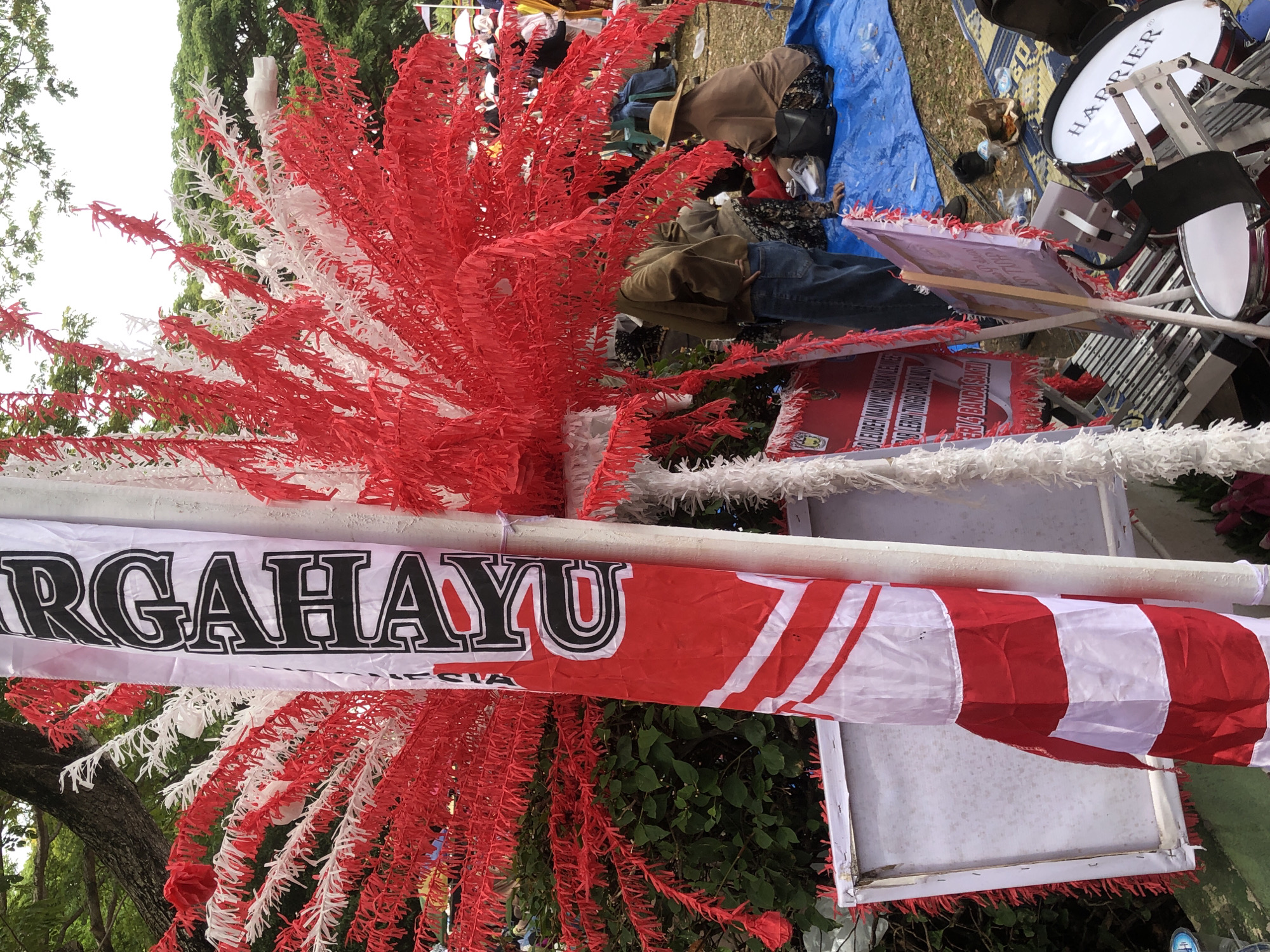 In Lhokseumawe, Indonesia, a close-up shot captures a decorative pole adorned with red and white streamers, possibly signifying national colors. A vertical banner, predominantly white with bold black text and red accents, hangs prominently. The text appears to be in Indonesian. In the background, a group of people are gathered, some seated on a blue tarp, while others stand. One person wears a brown jacket and blue jeans, while another is seated and wearing a straw hat and brown clothing. The environment includes trees and foliage, suggesting an outdoor setting. In the foreground, musical instruments, including drums, are visible.