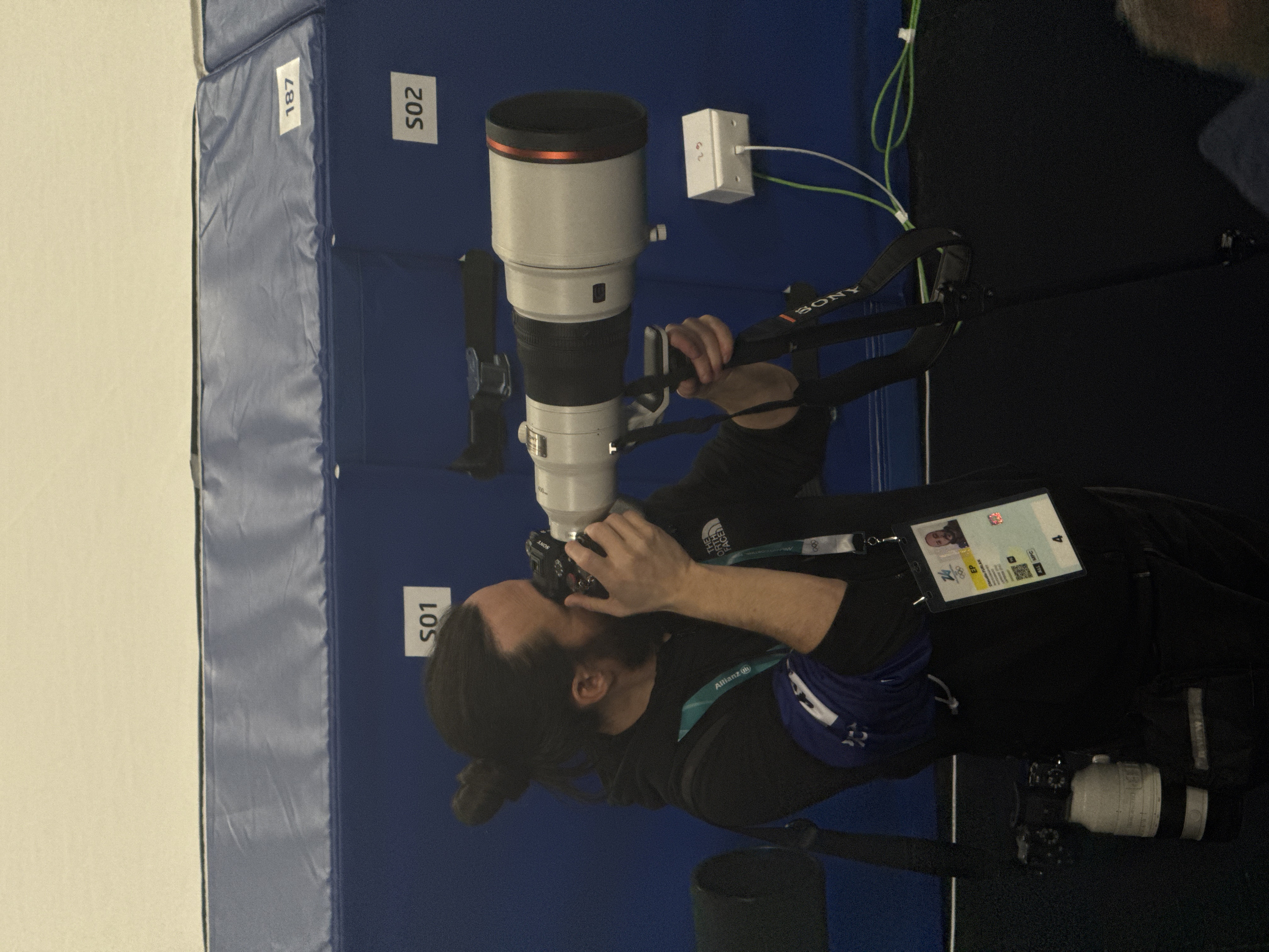 An indoor scene in Rho, Italy, features a male photographer operating professional camera equipment. The individual, positioned off-center left, has dark hair tied in a bun, a beard, and wears a black long-sleeve shirt with "THE NORTH FACE" logo. An event credential hangs from his neck, displaying his photo, the name "Claudio FURLAN," the Olympic rings, "MILANO CORTINA 2026," and an "Allianz" logo. A blue armband is present on his left arm. He is looking through the viewfinder or at the screen of a black Sony mirrorless camera, which is fitted with a large white telephoto lens featuring a red front ring. This camera and lens assembly is mounted on a black tripod with "SONY" branding.
The background consists of blue padded panels, upon which white labels bearing the alphanumeric codes "S01," "S02," and "187" are affixed. A white electrical box, marked "23" on its top surface, is mounted on the blue wall to the right, with a white and a green cable plugged into it, and additional green cables coiled on the dark floor below. A black strap is visible hanging on the blue wall behind the photographer's head. A partial view of a white cylinder with black straps is present on the far left. The upper background is a plain, light-colored surface. A blurry portion of another individual's head is visible in the lower right corner of the frame.