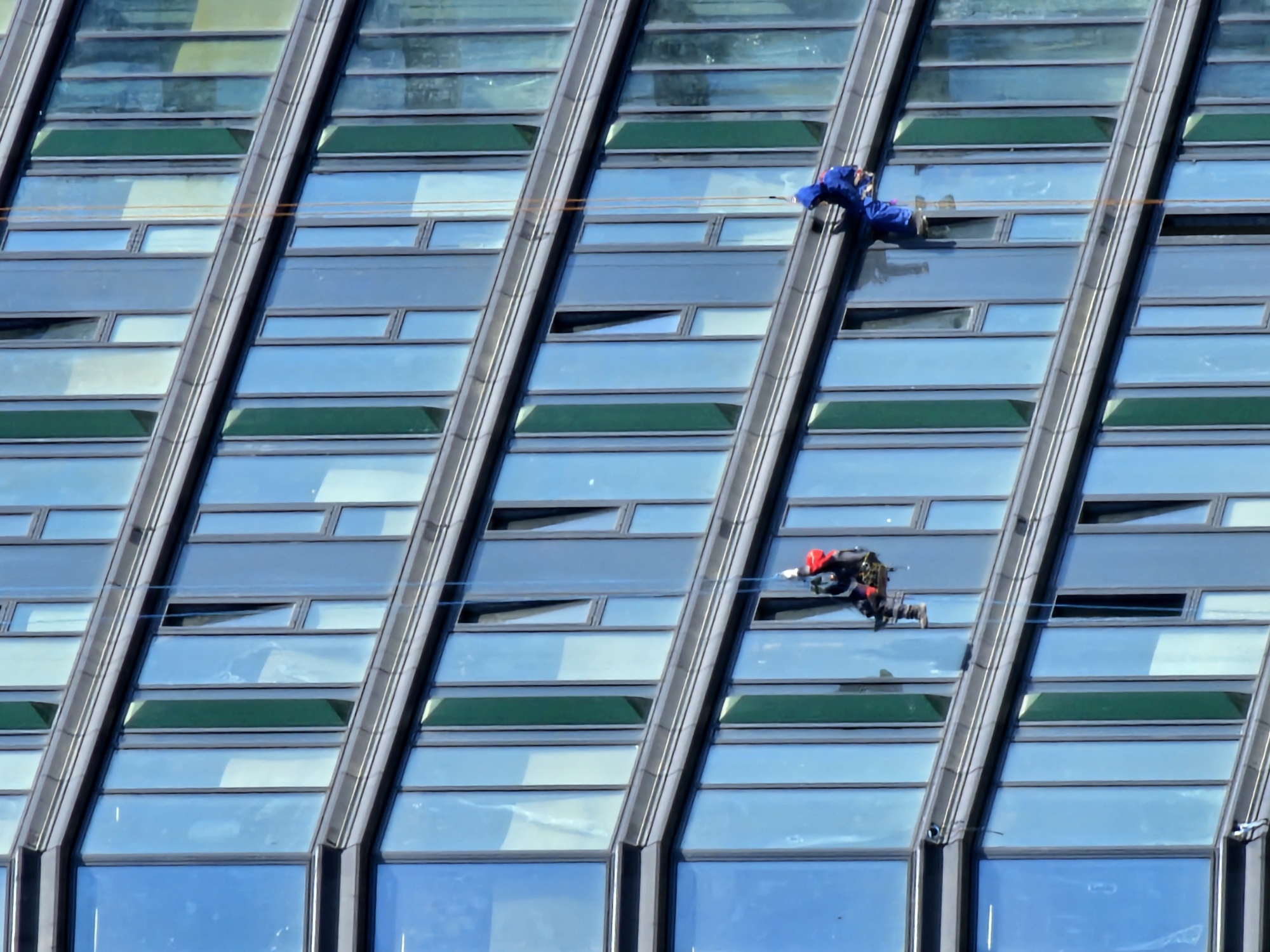 Two window cleaners are rappelling down the side of a modern glass-fronted building. The building has multiple floors with a pattern of alternating blue and green tinted windows, separated by dark gray horizontal frames. The cleaner on the left is wearing a red jacket and dark pants, suspended by ropes attached to a harness. The cleaner on the right is dressed in a blue jumpsuit, also secured by ropes. The scene takes place in Moscow, Russia, indicated by the architecture and urban setting.