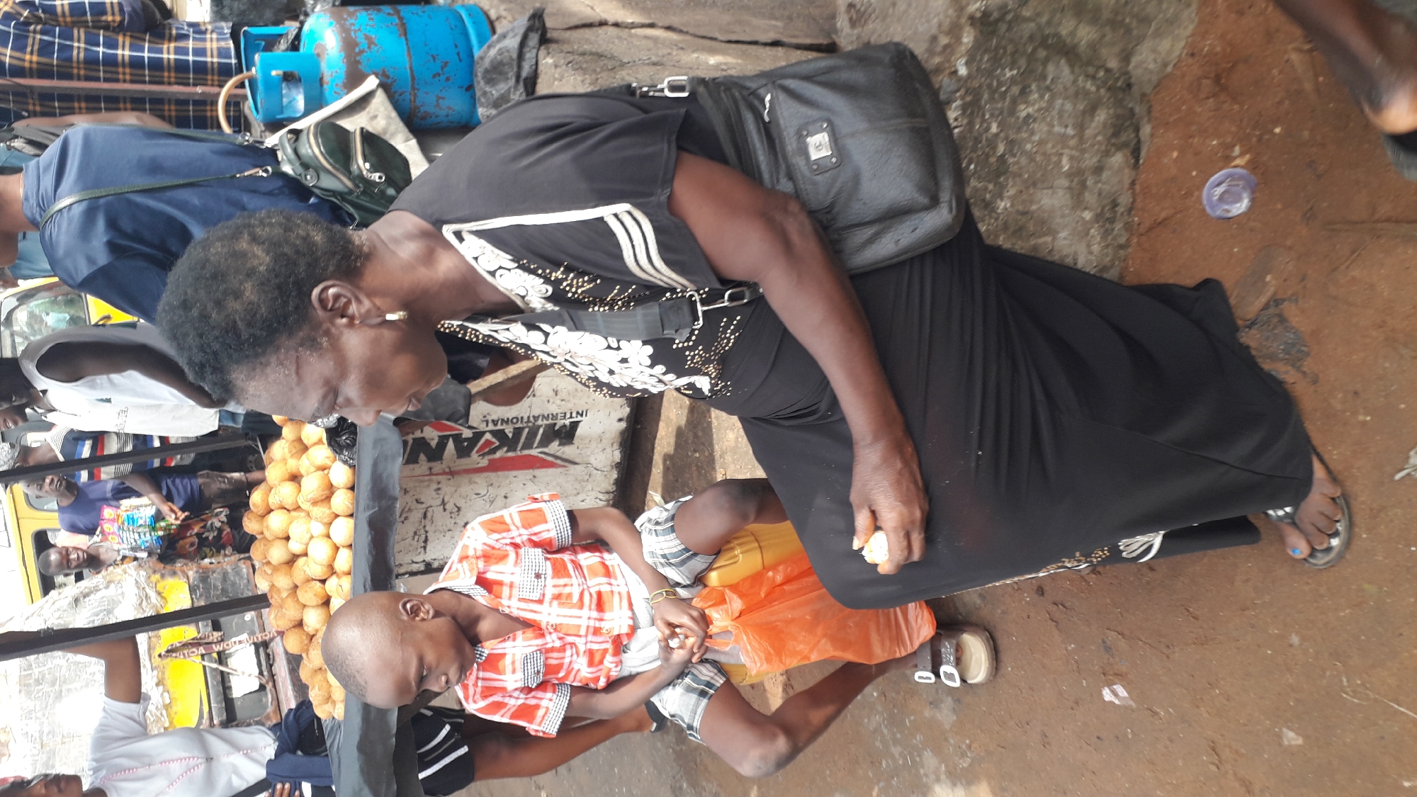A medium shot shows a woman and a boy sitting outdoors in Nkpor, Nigeria. The woman, who is in the foreground and appears to be in her late 40s, is wearing a long black dress with white floral patterns and a black handbag on her right hip. She is looking to her left, and her right hand is holding a small, white object, possibly food. The boy, who is to the woman's left and appears to be around 7 years old, is wearing a red and white checkered shirt and plaid shorts. He is looking down and to his left, with an orange plastic bag at his feet. Behind them, there is a table laden with a pile of round, golden-brown objects, likely food items such as plantain balls or puff puff, with a blue propane tank visible to the right. Other people and parts of a yellow vehicle are visible in the background, suggesting a market or roadside stall setting. The ground is made of reddish-brown earth, with some scattered debris.