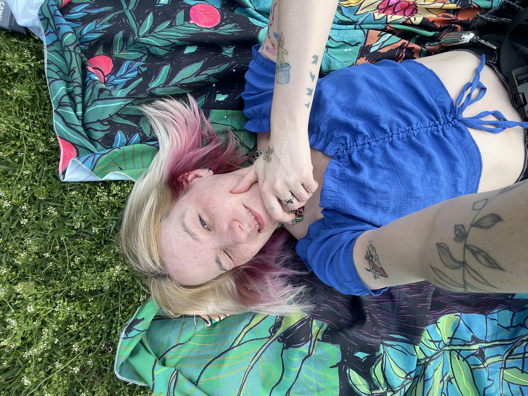 A person is lying on a colorful blanket on grassy ground, possibly in a park in Palilula Urban Municipality, Serbia. The person has blonde and pink hair, freckles, and tattoos on their arms and hands. They are wearing a blue top. The blanket has a vibrant, tropical-themed pattern with flowers and leaves. The grass is speckled with small white flowers.