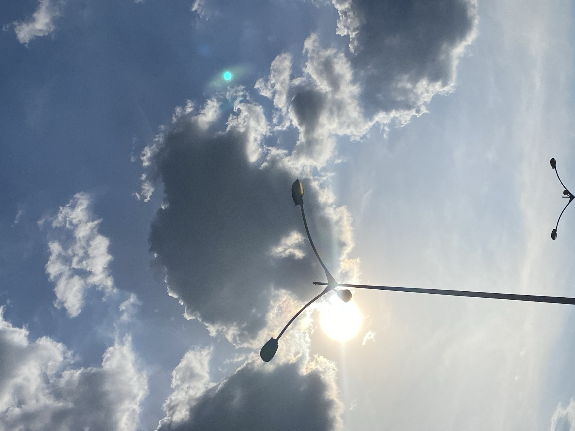 The image displays a daytime sky over the city of Unknown, Unknown, characterized by a mix of blue sky and dense cumulus cloud formations. The sun, appearing as an intense, overexposed white circle, is positioned in the lower-left quadrant, partially obscured by a cloud and a prominent streetlight. Distinct sun rays emanate from behind the clouds. A tall, dark-silhouetted streetlight with two curved arms and fixtures is centrally located, with one fixture directly in front of the sun. A second, smaller streetlight is partially visible in the bottom-right corner, also silhouetted. A bright, circular, turquoise-green lens flare is present in the upper-right portion of the frame. No individuals, specific actions, or additional objects are depicted in the scene beyond the atmospheric conditions and the two utility poles.