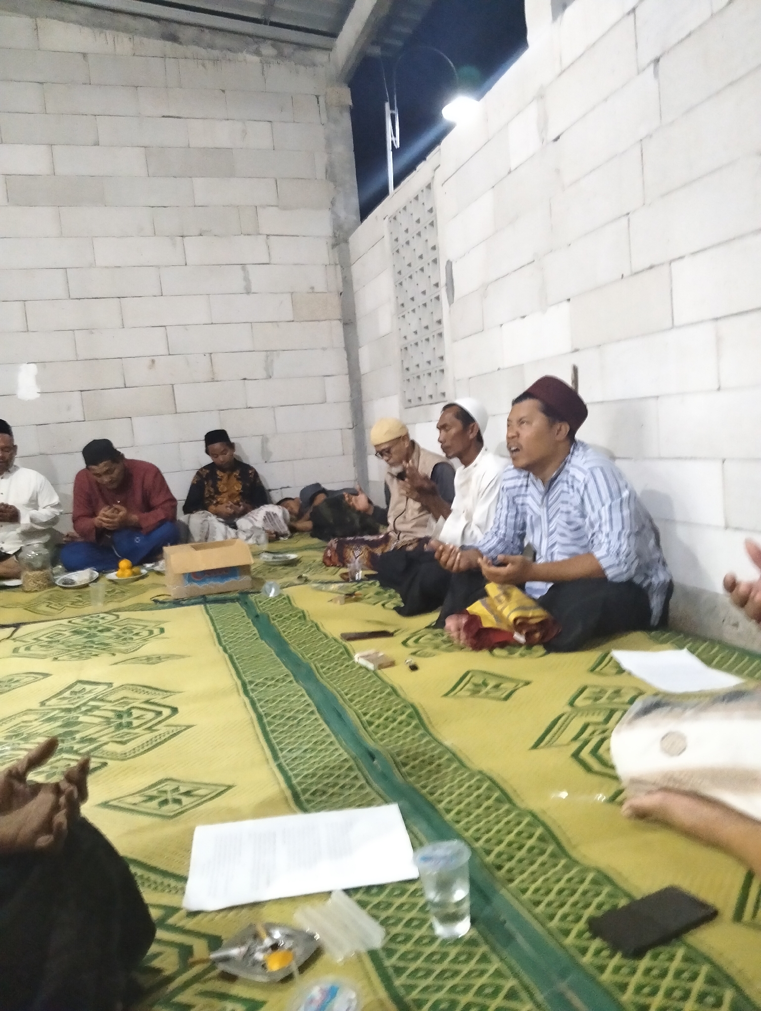 The image shows a group of men seated on a yellow and green patterned mat, likely in a religious gathering or meeting. The men are all wearing traditional attire, including peci hats and long-sleeved shirts. Some men are engaged in prayer or contemplation, with their hands clasped. There are several objects on the mat, including a glass of water, a mobile phone, papers, and a small box. The background consists of a white cinder block wall, suggesting an indoor or semi-outdoor setting. The lighting is dim, with some artificial light sources visible. The scene appears to be in Gedangan, Indonesia, based on the context provided.
