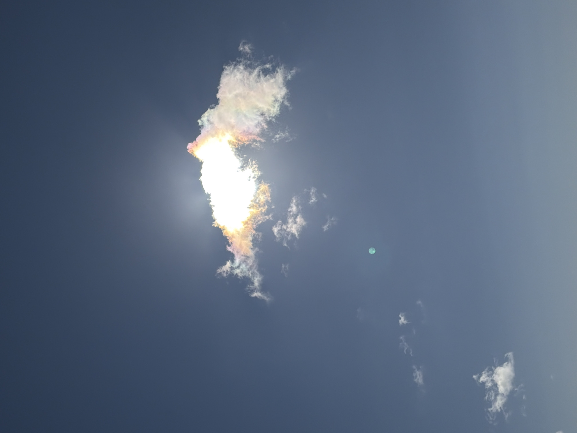 This is a photograph of a cloudy sky above Ozerskoe, Russia. The sun is shining brightly behind a large, white cloud, creating a halo effect. The cloud itself has iridescent colors, suggesting it is a nacreous cloud, also known as a mother-of-pearl cloud. These clouds are rare and are typically seen at high altitudes. There are several smaller, wispy clouds scattered across the sky. A faint, circular light artifact is visible in the lower right quadrant of the image, which may be a lens flare or reflection. The sky is a clear, deep blue. There are no people or man-made structures visible in the photograph.