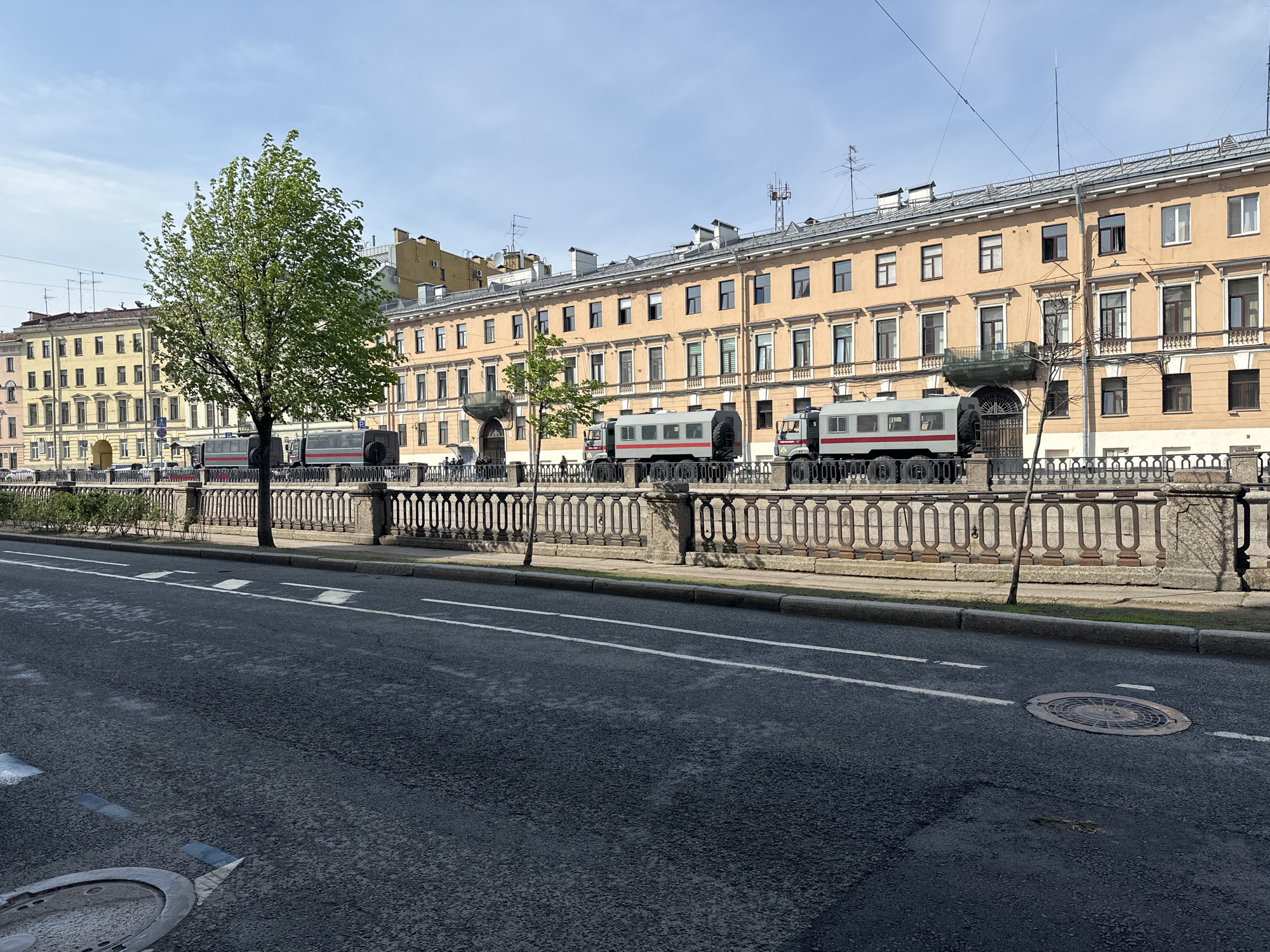 The image depicts a street scene in Saint Petersburg, Russia. Along the embankment, there are several specialized vehicles, likely for law enforcement or security purposes, parked behind an ornate stone railing. A young green tree stands prominently to the left. Adjacent to the embankment is a long, multi-story building with a beige facade and numerous windows. The street is asphalt, showing typical wear and tear, and includes painted lane markings and a manhole cover. The sky is clear and blue.