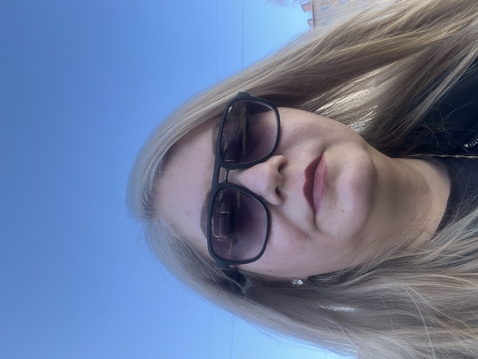 A fair-skinned woman with long blonde hair and dark lipstick is pictured wearing sunglasses against a blue sky. The reflection in her sunglasses reveals a city setting, possibly Glazov, Russia. A building is partially visible in the upper right corner of the sunglasses' reflection. A small earring is visible on the woman's ear. The woman appears to be wearing a black top or jacket.