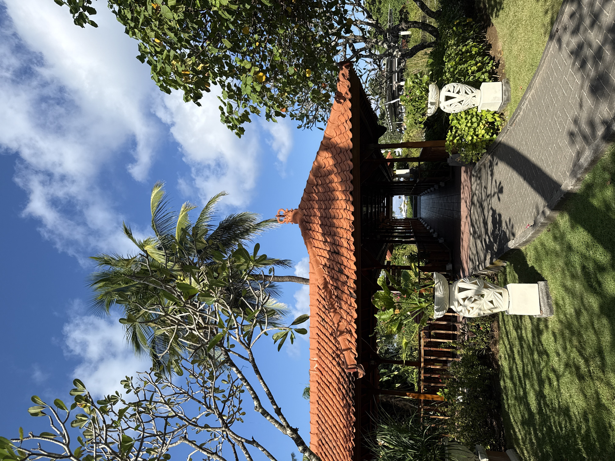 This image captures a bright, serene outdoor scene, likely a resort garden or traditional compound in Kuta Selatan, Indonesia, viewed as if in a landscape orientation.
The central subject is a traditional Indonesian-style pavilion or building, characterized by its distinctive reddish-orange, layered tiled roof. A decorative finial, possibly depicting a mythical creature or traditional motif, adorns the apex of the roof. The structure beneath the roof appears to be made of dark wood, suggesting a sheltered pathway or open-air common area.
The setting is lush and tropical. To the left of the building, a tall palm tree with vibrant green fronds stands out against the blue sky, alongside another tree with light-colored, gnarled branches and scattered green leaves, possibly a frangipani. Various other leafy green trees and shrubs frame the scene. The ground is covered with neatly manicured green grass on the right, where strong sunlight casts distinct shadows, indicating a bright late morning or early afternoon.
A stone-paved pathway curves from the right foreground towards the entrance of the building. Flanking this path, near the building, are two ornate, intricately carved white stone sculptures, resembling decorative urns or pedestals. The sky above is a clear, bright blue with scattered white, wispy clouds.
No specific activity or event is taking place; the scene is static and peaceful. The traditional architecture, tropical foliage, and clear, sunny weather strongly convey the location's Indonesian character. There is no visible text in the image.