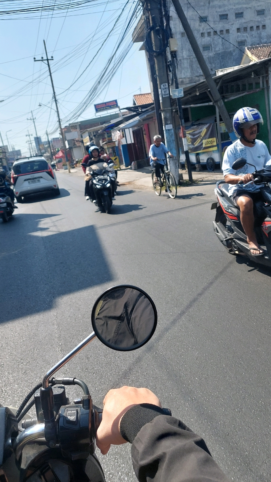 This is a first-person perspective, low-angle medium shot of a person riding a motorcycle on a sunny day on a paved road in Sidoarjo, Indonesia. The road has lane markings and shadows from overhead wires and surrounding buildings. Traffic includes other motorcycles, a white car, and a person riding a bicycle. The rider's hand, wearing a black jacket, is visible on the handlebars, which include controls and a rearview mirror reflecting the sky and overhead wires. In the background, commercial buildings with signs and utility poles with a dense network of electrical wires line the street. The overall scene depicts a typical urban street environment with active traffic.