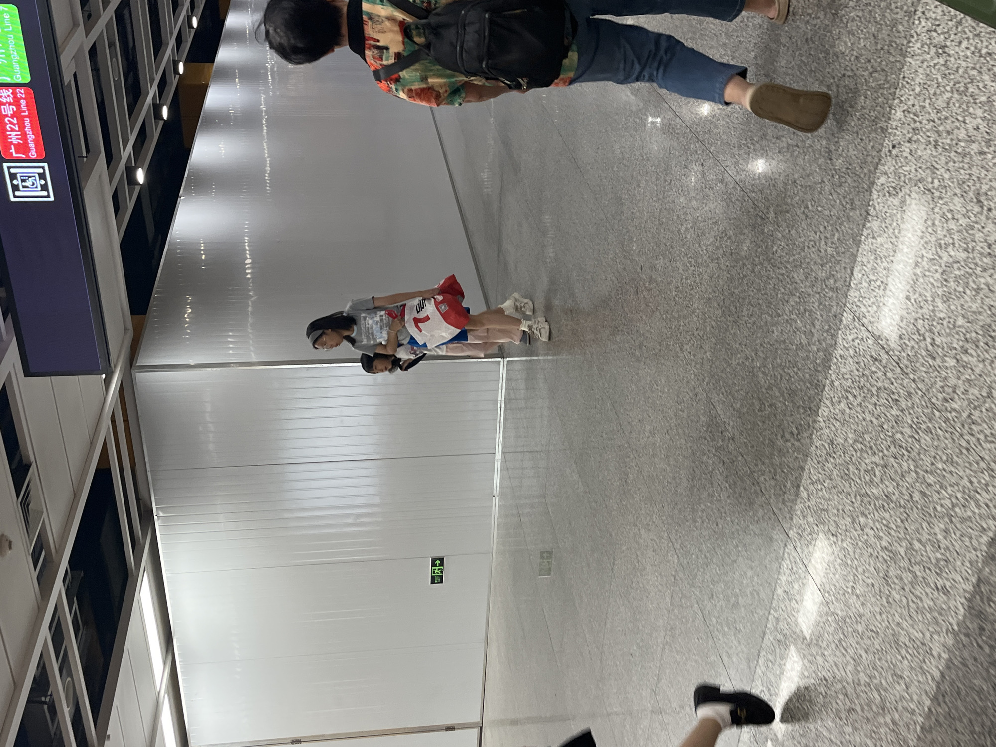 This is a photograph taken from a low angle inside a modern building, likely a subway station in Panyu District, China. The floor is a speckled gray tile with reflective qualities, showing reflections of the ceiling and overhead lighting. A large, white, corrugated wall dominates the background, stretching horizontally across the frame. On this wall, to the left, is a small green exit sign with an arrow pointing to the right.
In the middle ground, three people are walking away from the camera. Closest to the camera on the left is a person wearing a black shoe and white socks, partially visible. To their right is a woman wearing glasses and a grey t-shirt, carrying a white and red bag. She is accompanied by a young child, also partially visible. To the right of the woman, a person wearing a colorful patterned shirt and a black backpack is walking away from the camera. They are wearing blue jeans and brown shoes.
In the upper part of the image, a sign is visible with Chinese characters and English text, indicating "Guangzhou Line 22" and "Guangzhou Line 7". The ceiling is made of a grid of white panels with recessed lighting. The overall impression is of a clean, well-lit interior public space.
