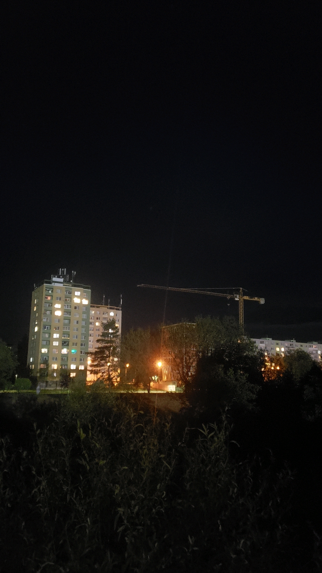 The image depicts a nighttime scene in the city of Prešov, Slovakia. In the foreground, there is a dense thicket of dark foliage. Behind the foliage, two tall, illuminated apartment buildings are visible. The building on the left is lighter in color and has numerous windows lit up. The building to its right is also illuminated. A large construction crane with a yellow boom stretches across the middle ground, extending from right to left, and is positioned between the apartment buildings and more distant structures. Further in the background, another row of illuminated buildings is partially obscured by trees. A street lamp casts a warm orange glow on the scene. The overall environment is urban and appears to be residential, with ongoing construction indicated by the presence of the crane.