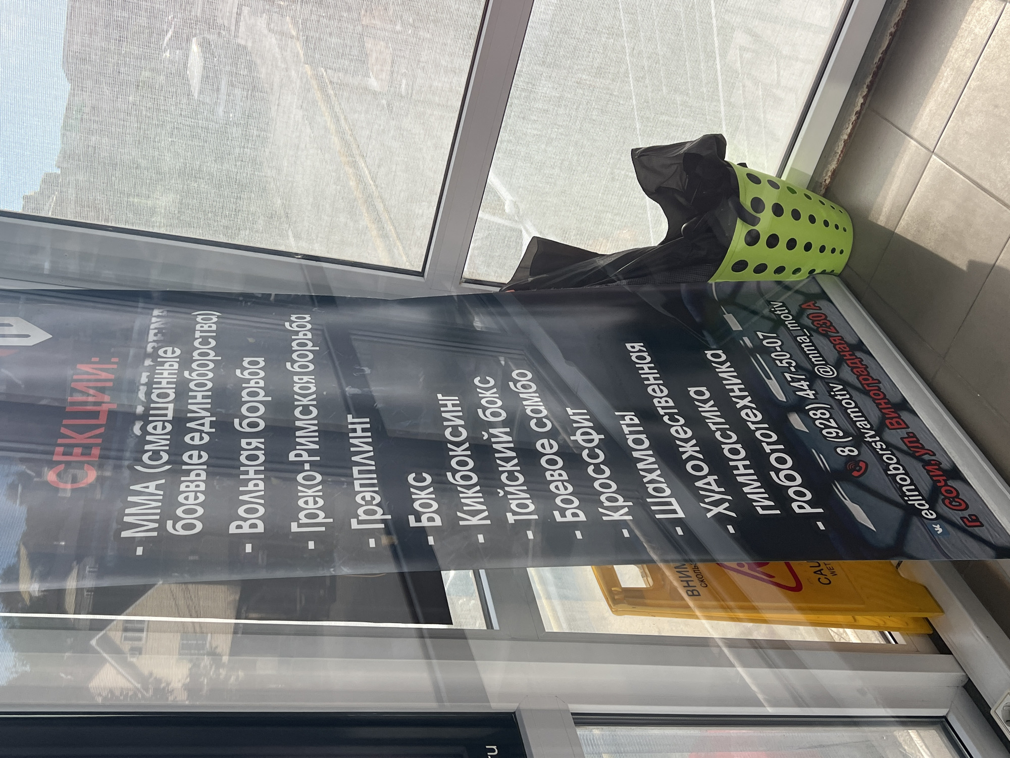 This media file presents an indoor scene, likely within a sports facility or gym, with an informational banner prominently displayed next to a large window. The image is oriented sideways.
The main subject is a black banner with white Russian text, advertising various sections or disciplines available. The heading reads "СЕКЦИИ:" (SECTIONS:), followed by a list of activities:
- MMA (смешанные боевые единоборства) - Mixed Martial Arts
- Вольная борьба - Freestyle wrestling
- Греко-Римская борьба - Greco-Roman wrestling
- Грэпплинг - Grappling
- Бокс - Boxing
- Кикбоксинг - Kickboxing
- Тайский бокс - Thai boxing
- Боевое самбо - Combat Sambo
- Кроссфит - CrossFit
- Шахматы - Chess
- Художественная гимнастика - Rhythmic gymnastics
- Робототехника - Robotics
Below the list, contact details are provided: a phone number "8 (928) 447-50-07", social media handles "@mma_motiv" and "edinoborstvamotiv", and the address: "Г. Сочи, ул. Виноградная 230А" (Sochi, Vinogradnaya Street 230A), confirming the location in Sochi, Russia.
The setting is bright, indicating daytime, with ample natural light coming through the large window. A faint reflection of a building, possibly a house with a pitched roof, is visible on the windowpane. Adjacent to the banner, on the tiled floor, is a bright green, perforated plastic basket holding a black bag. Partially visible outside the window is a yellow "Caution Wet Floor" sign, with the English "CAU WET" and Russian "ВНИМ" (Attention) and "СКОЛЬ" (Slippery) discernable. No people are present, and no particular activity or event is taking place other than the display of this informational banner.